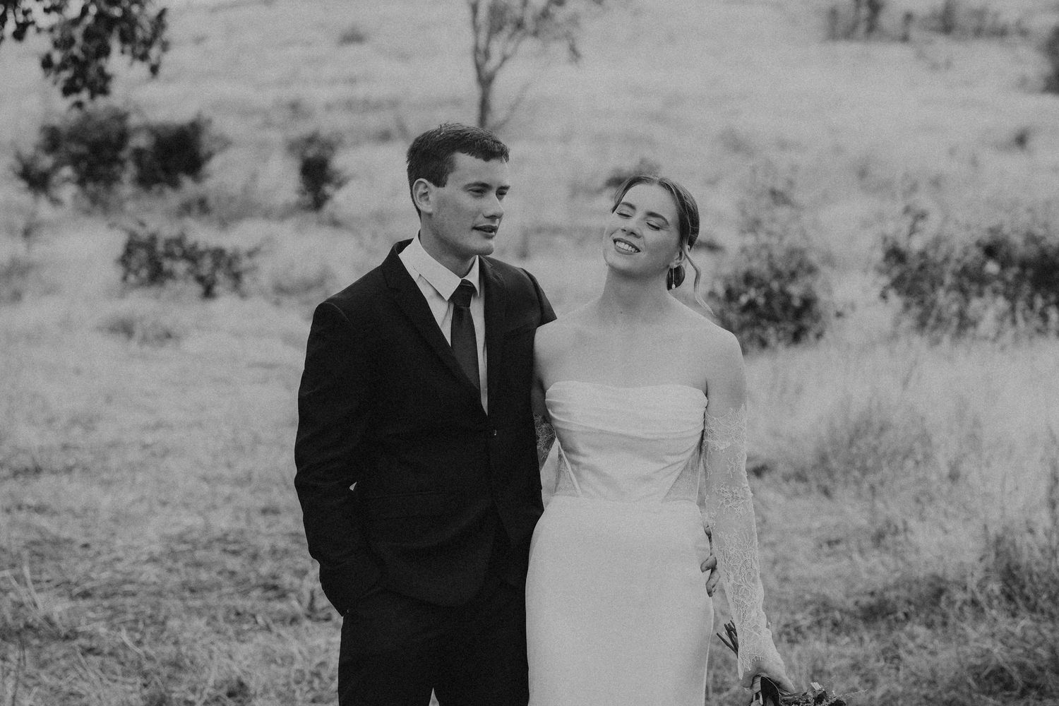 Black and white photo of a smiling bride and groom standing together in a field on their wedding day.