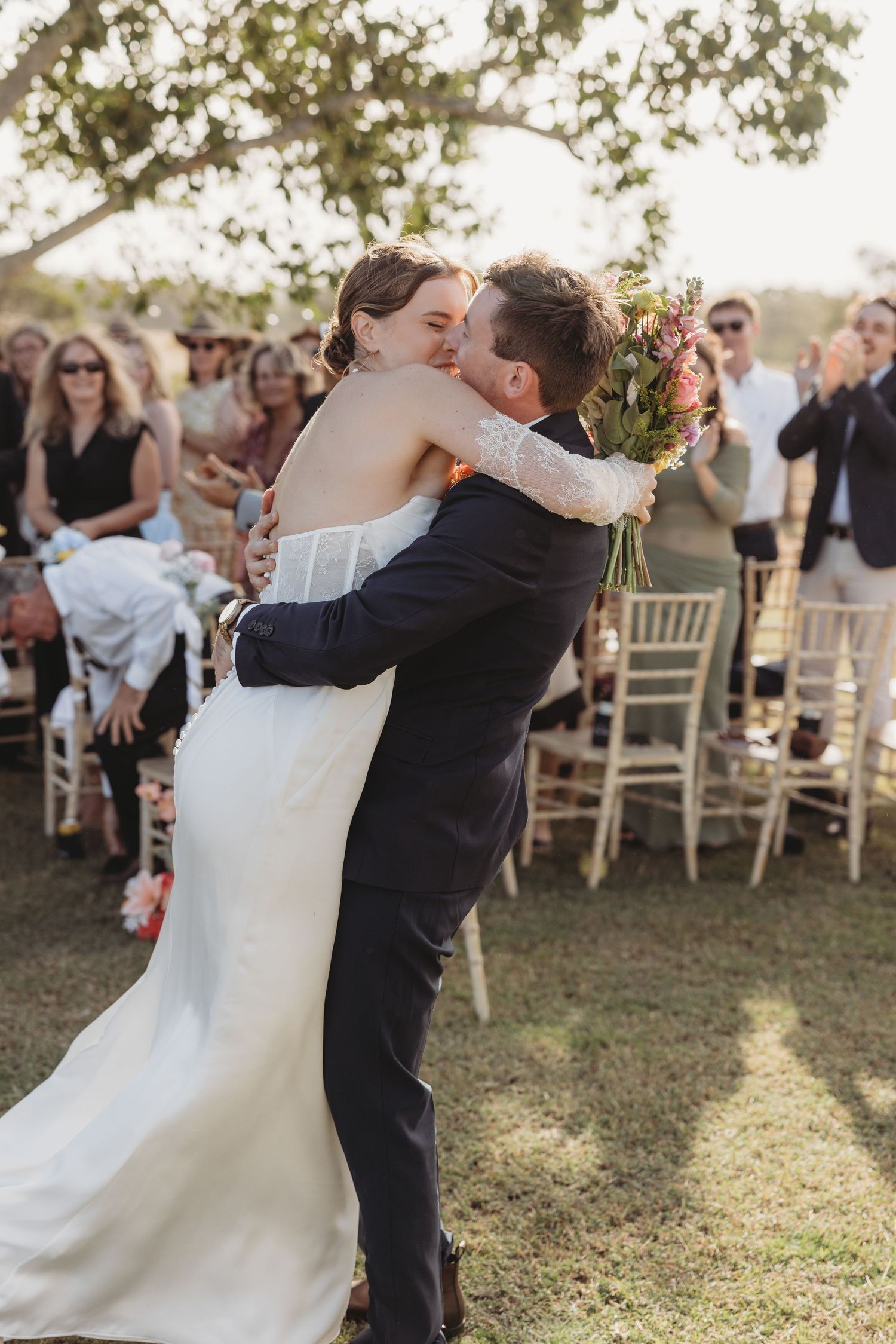 A groom lifts and kisses his bride in a white dress as wedding guests cheer at an outdoor ceremony.