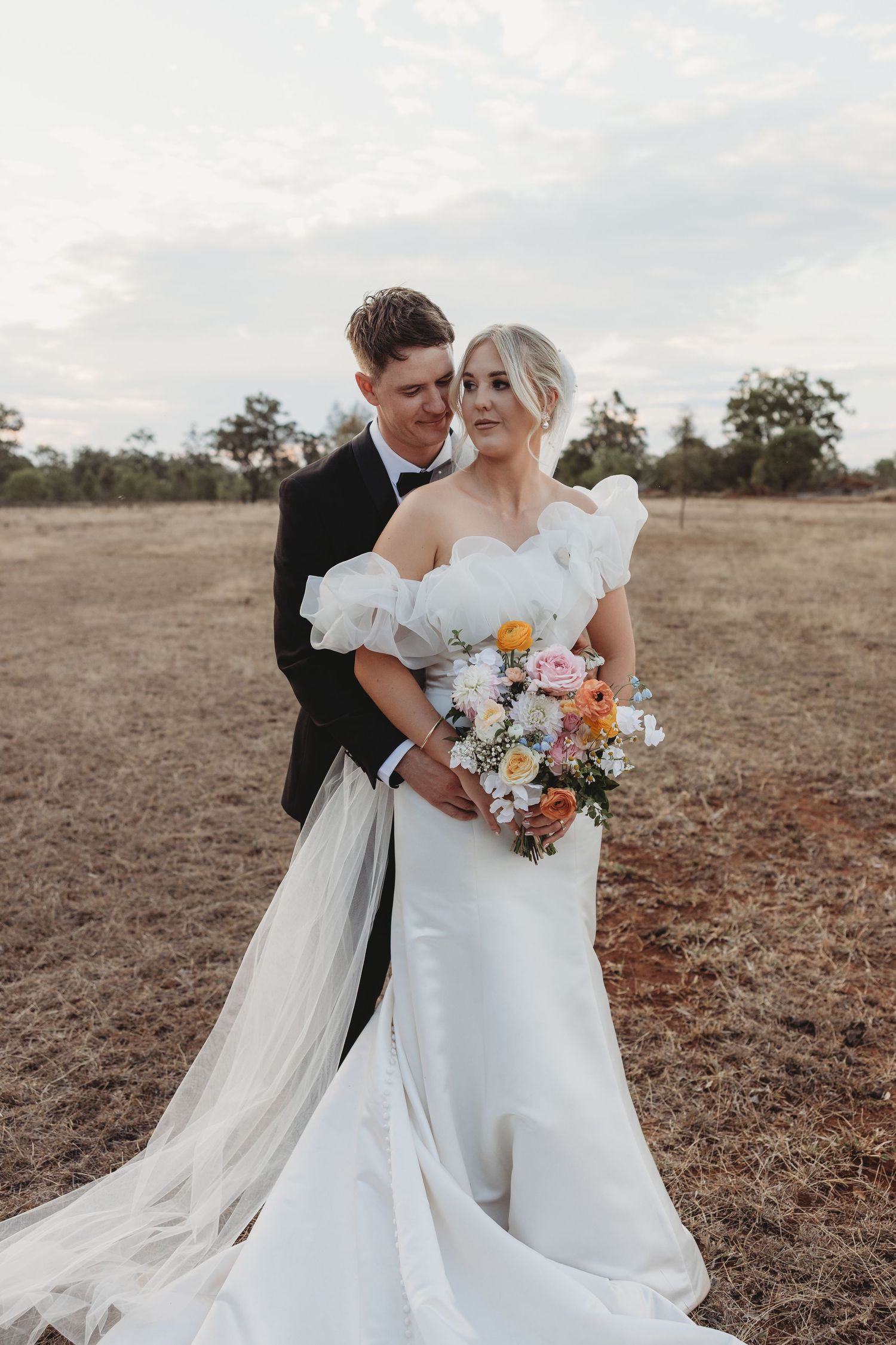 Newlyweds pose in rustic field with colorful bouquet and elegant white wedding dress.