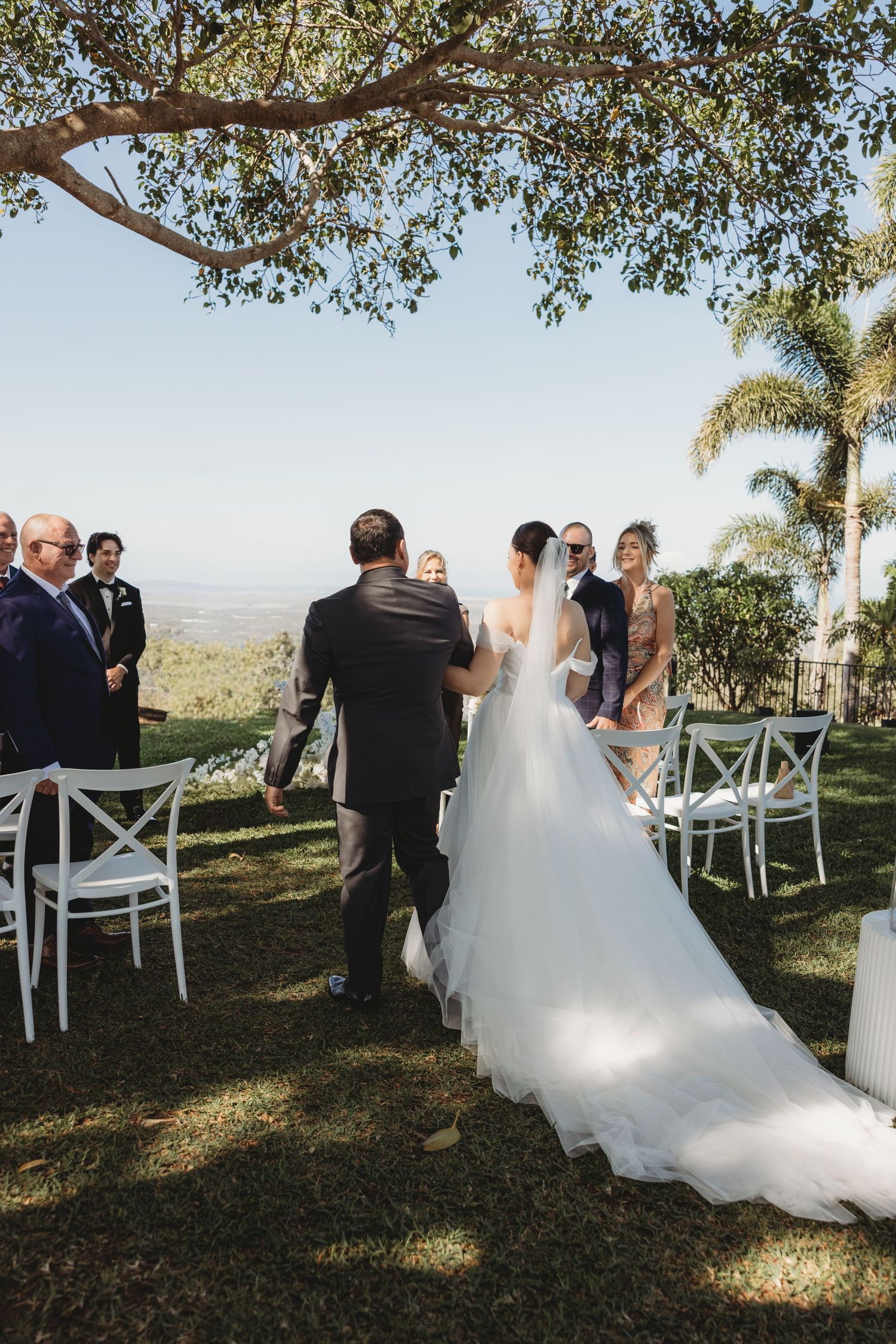 A bride in white gown walking down the aisle during an outdoor hilltop wedding ceremony.