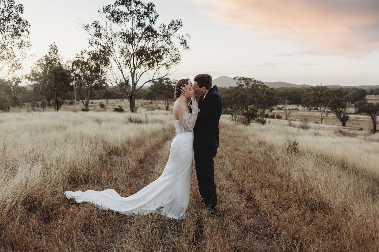 Bride and groom share romantic kiss in golden wheat field at stunning sunset wedding.