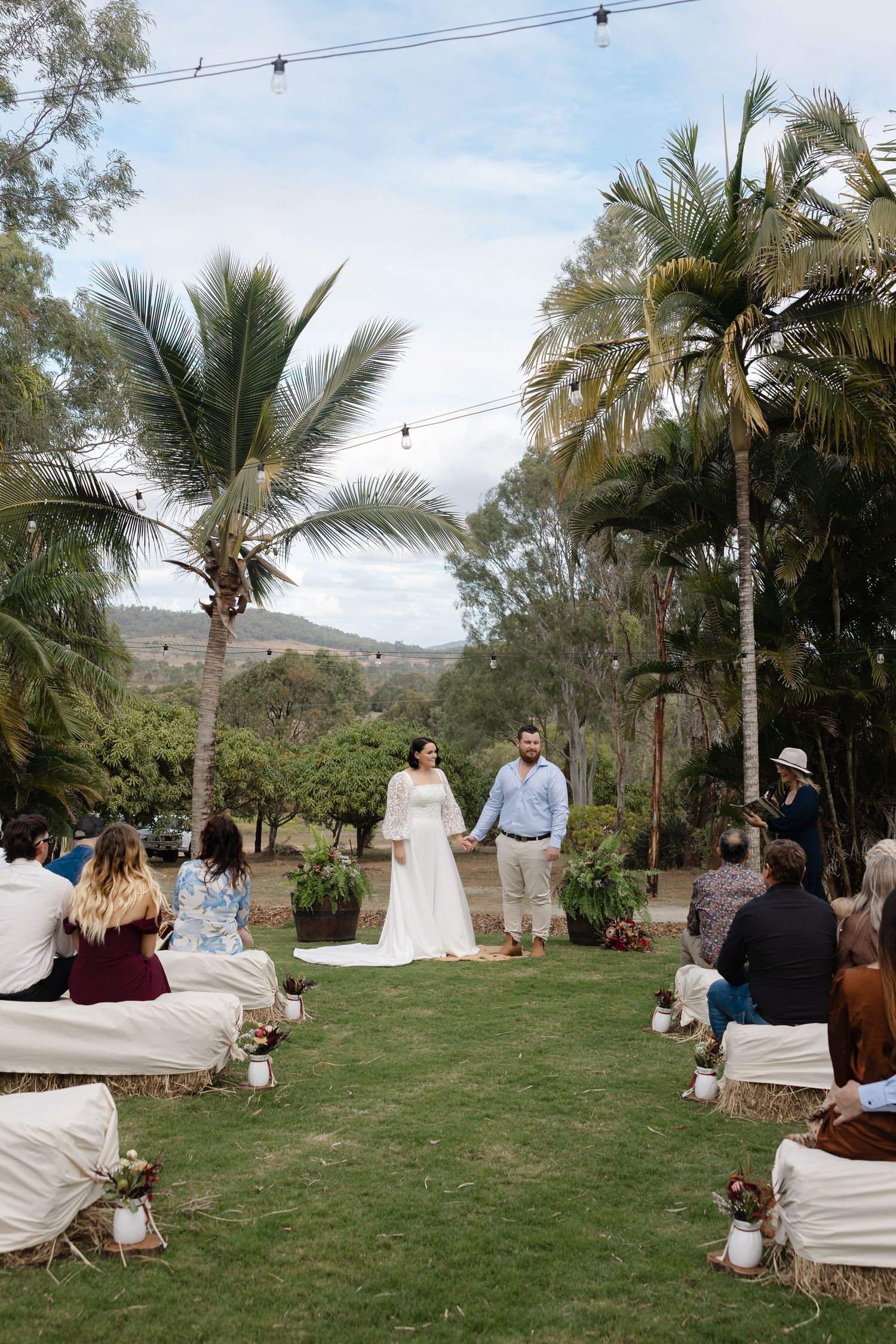 Outdoor wedding ceremony on green lawn with palm trees, couple holding hands at altar, guests seated on white cushions.