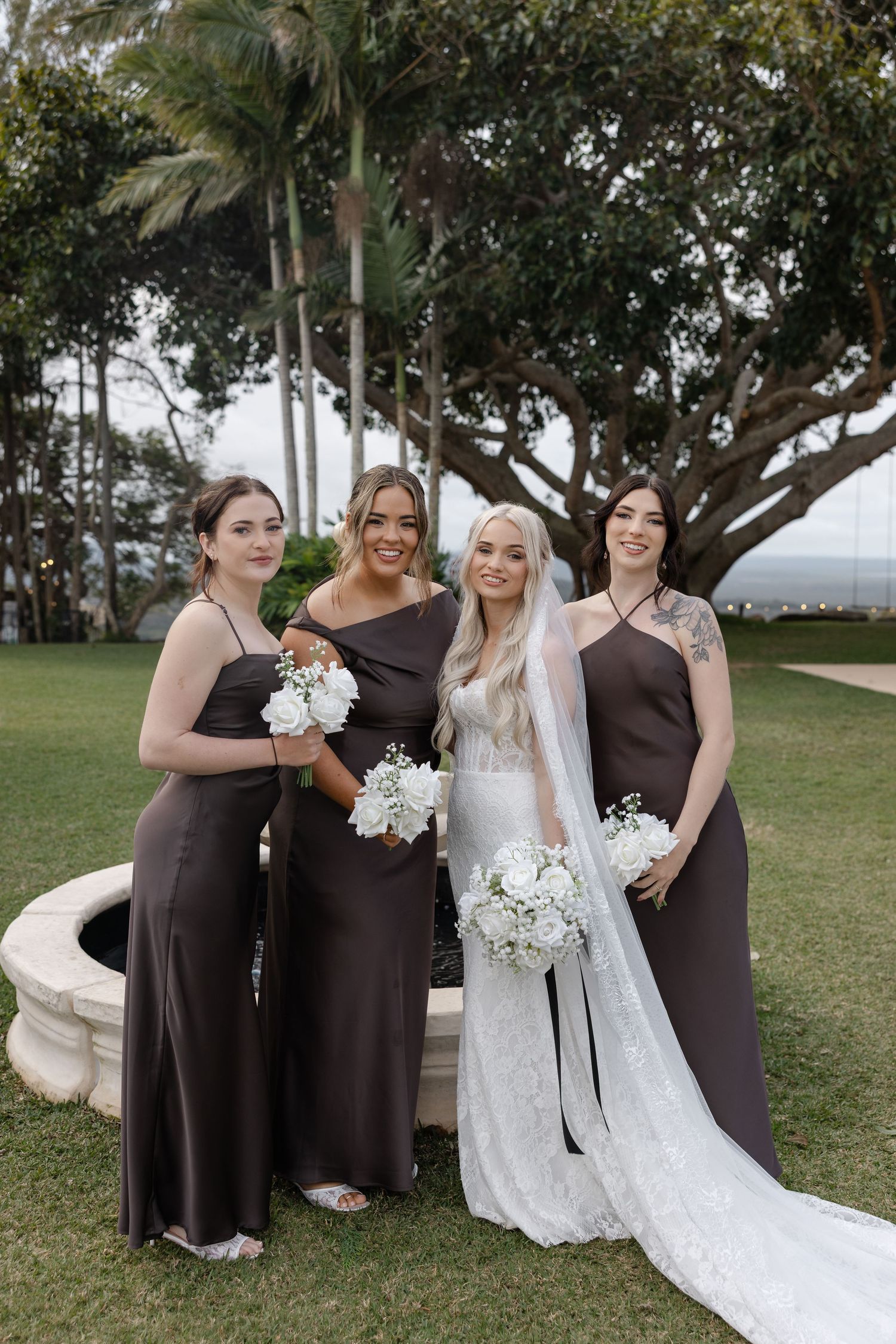 Bride in white lace gown with veil poses with three bridesmaids in brown satin dresses holding white bouquets outdoors.