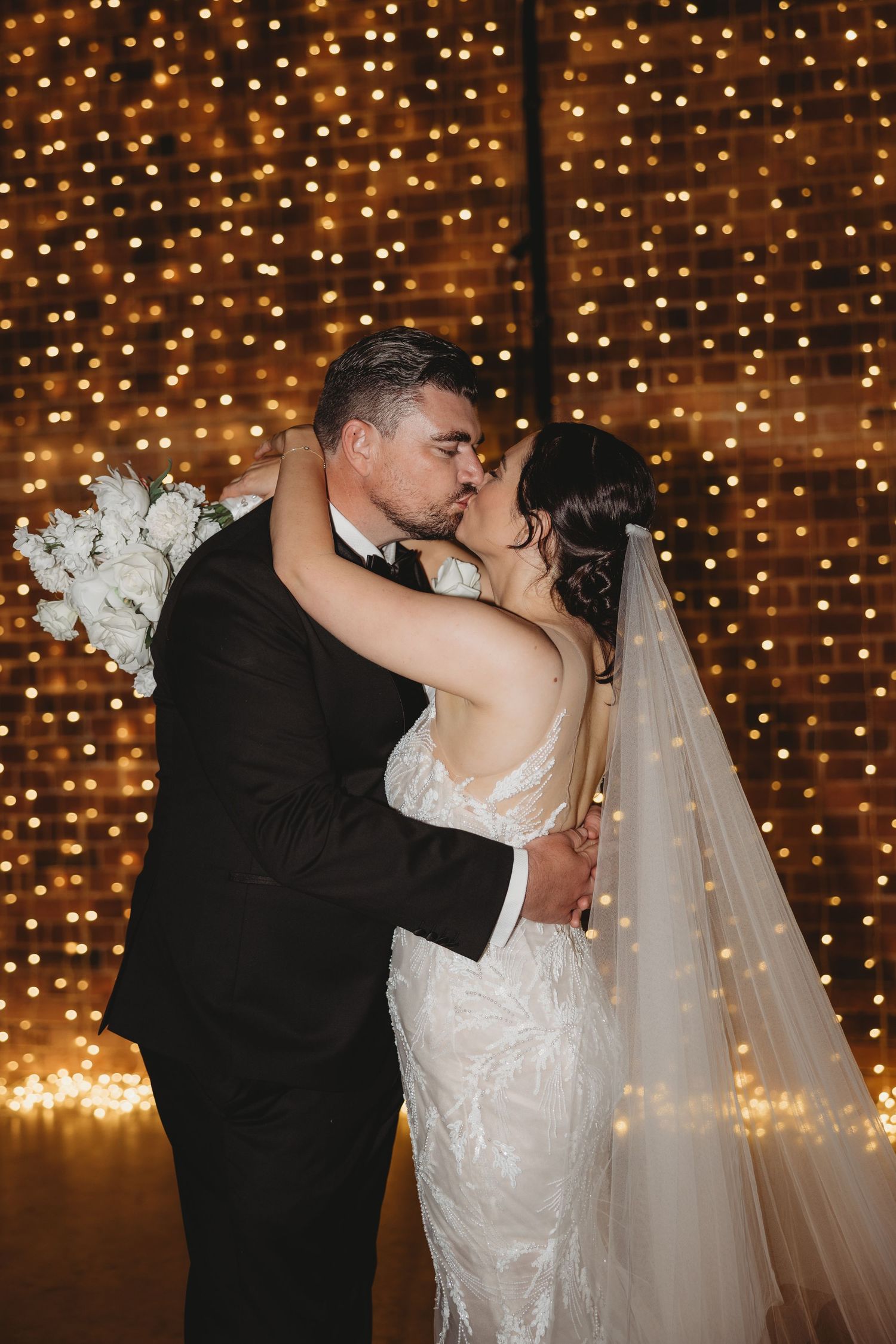 Romantic wedding couple sharing a kiss against a backdrop of twinkling string lights in a dimly lit venue.