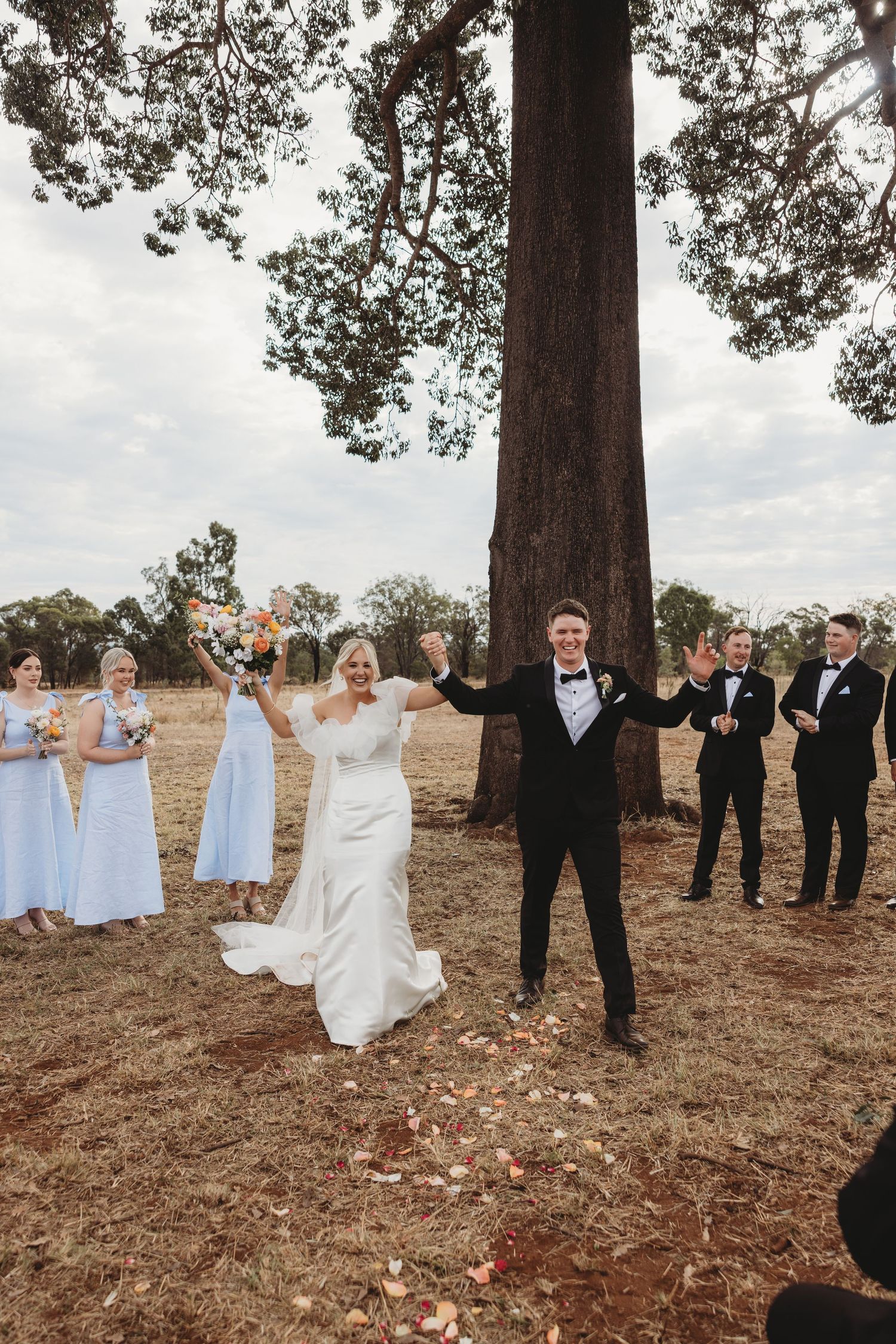 Wedding party arranged in casual poses under pine trees on hillside during outdoor celebration.