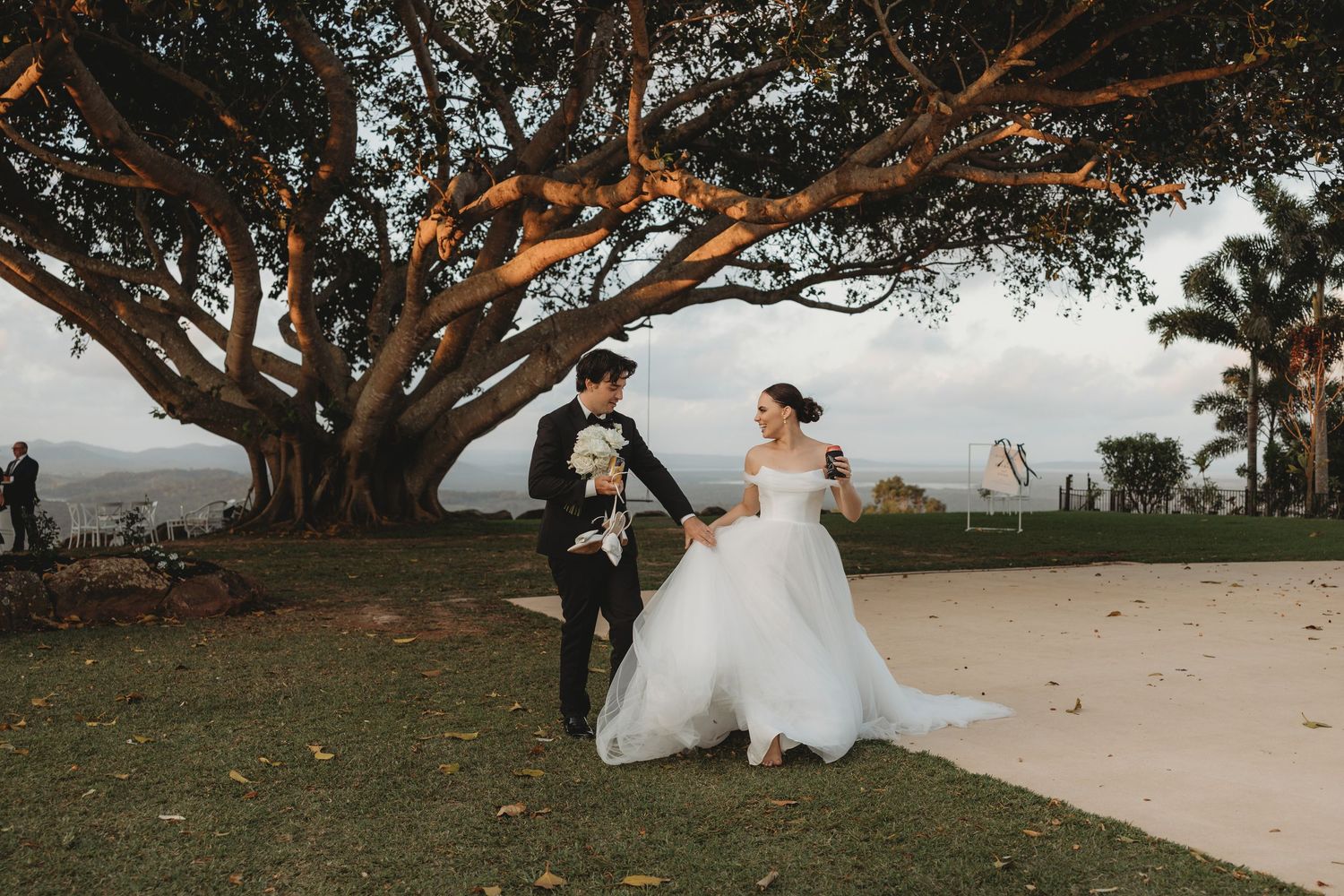 Wedding couple poses playfully under large tree at sunset.
