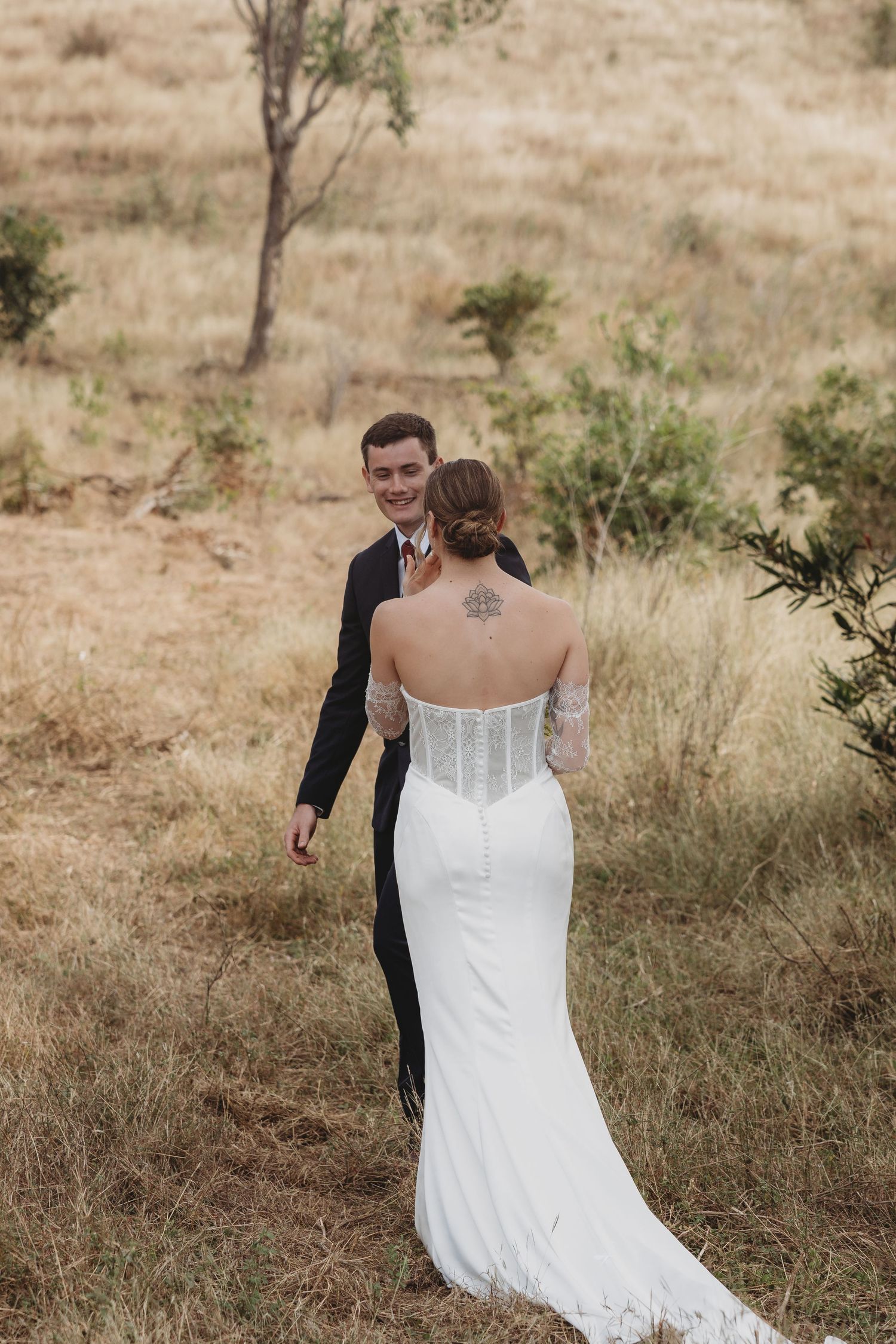 Bride in fitted white lace gown with back tattoo walks toward smiling groom in field.