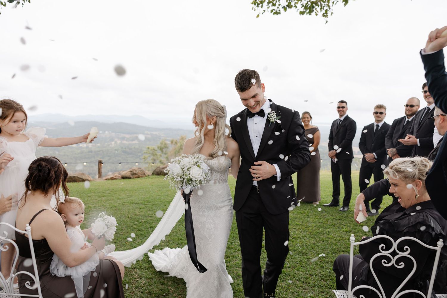 Bride and groom walk down the aisle as guests toss white confetti at an outdoor wedding ceremony.