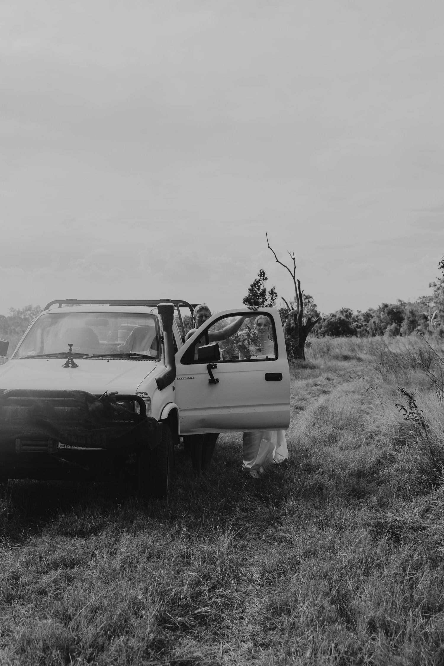 Black and white photo of a bride in a white dress standing beside a vintage truck in a rural field.