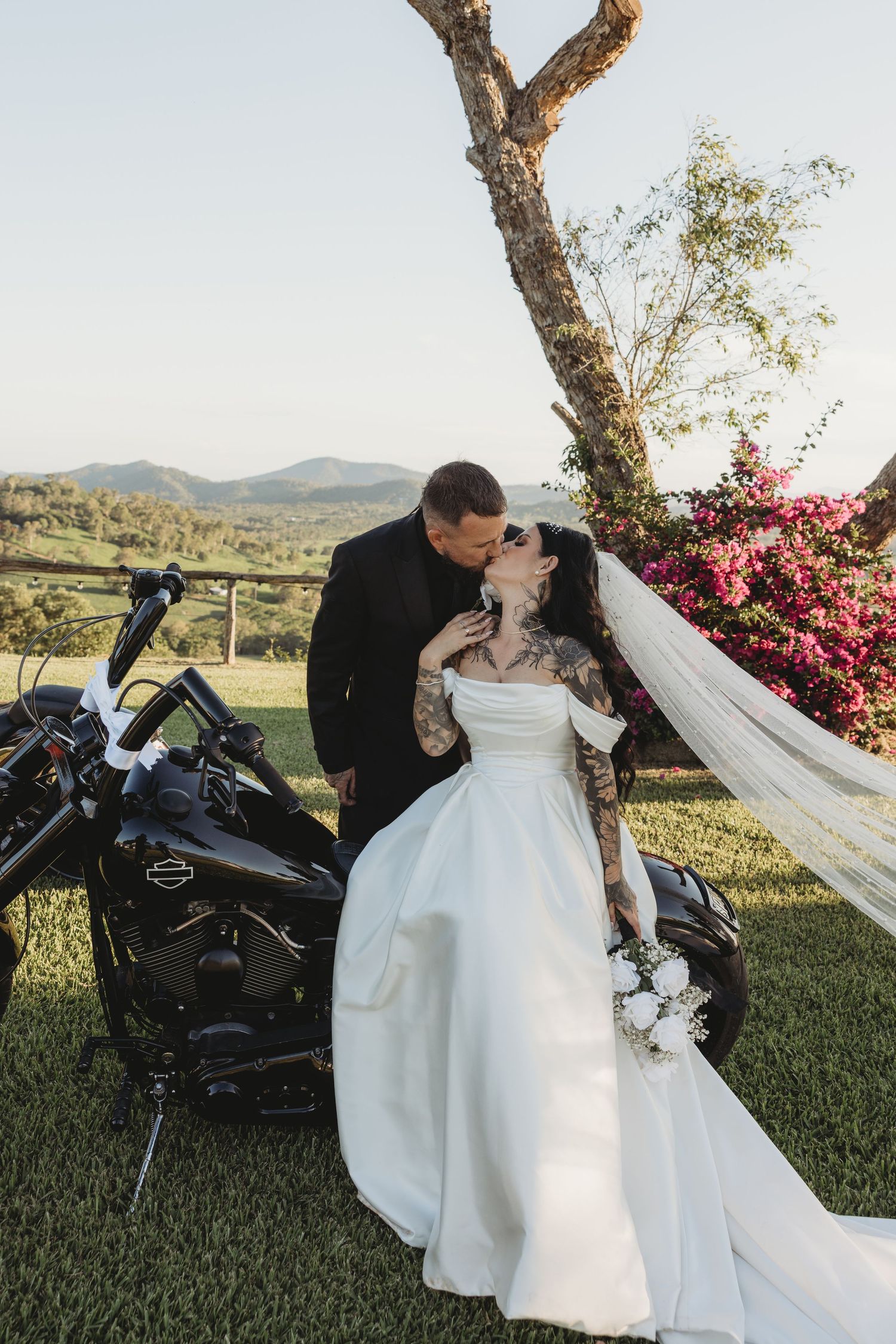 A romantic wedding portrait with a motorcycle, flowering tree, and rolling hills in the background.