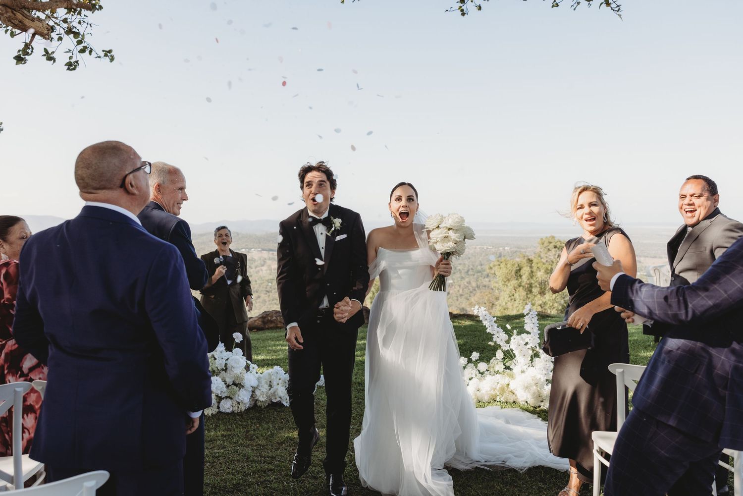 Wedding guests gather under a tree for an outdoor ceremony with white chairs and scenic views.