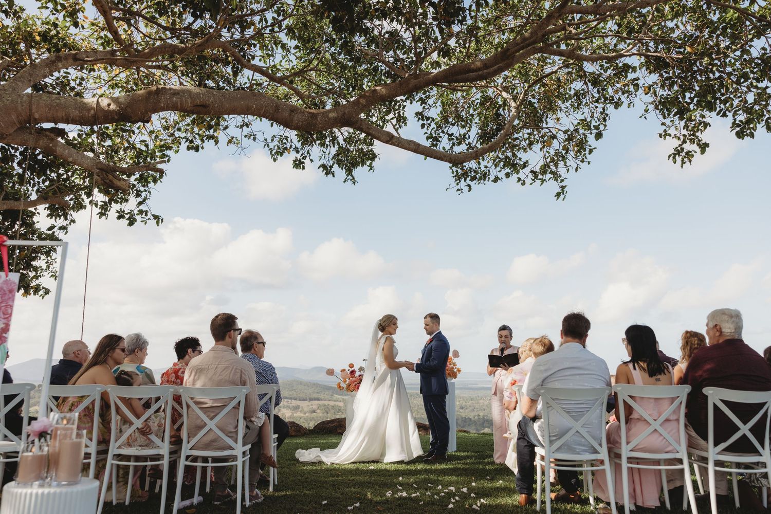 Outdoor wedding ceremony setup under a large tree with ocean views and white chairs.
