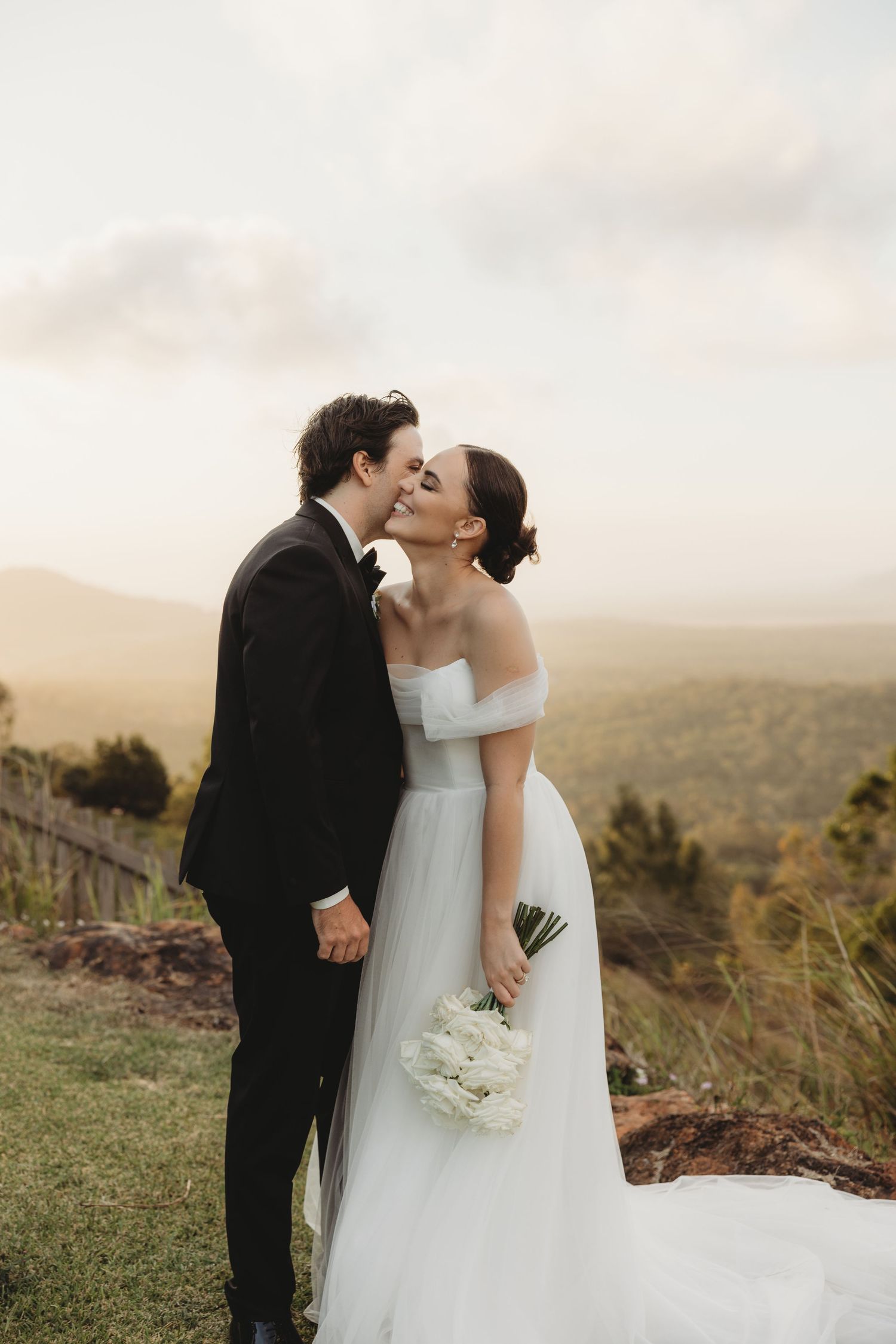 Newlyweds share a romantic kiss against a scenic hilltop backdrop at sunset.