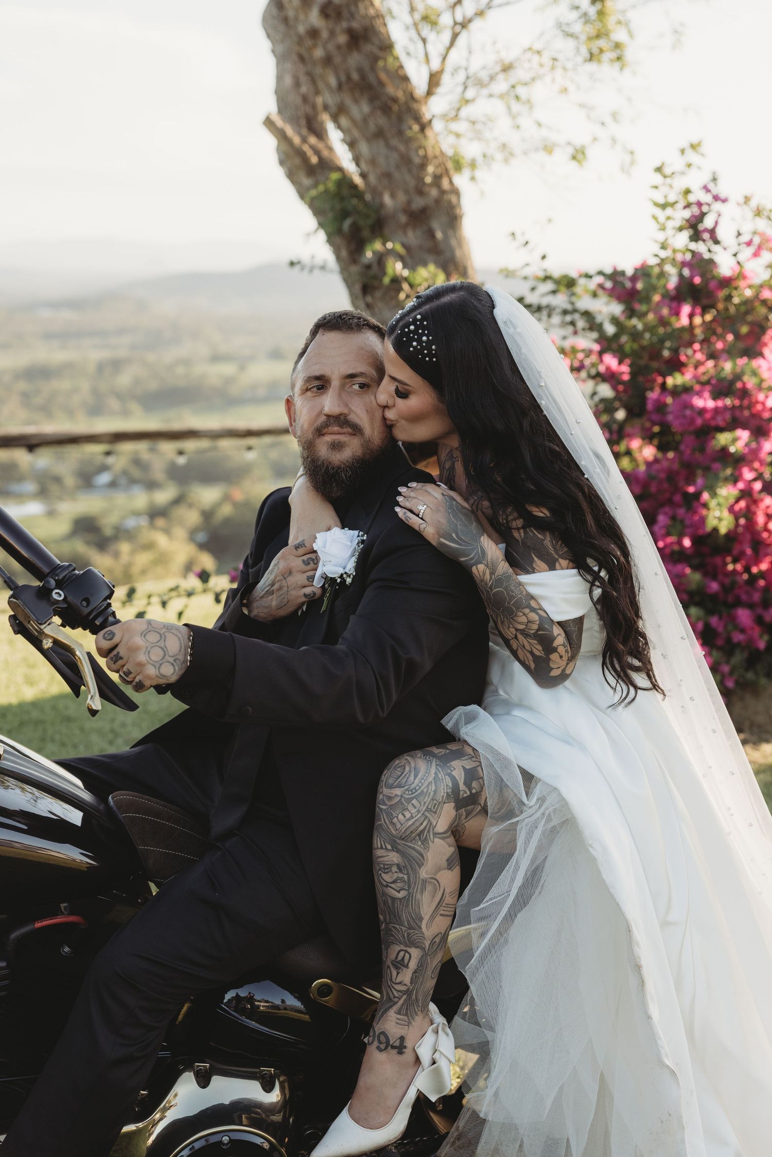 Wedding couple sharing intimate moment near motorcycle with scenic countryside backdrop.