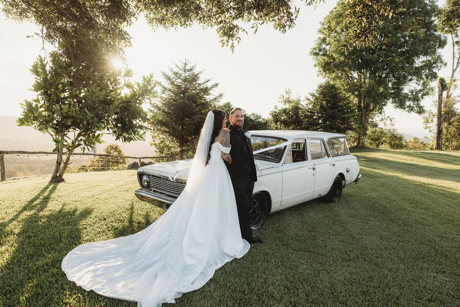 A vintage white station wagon serves as a backdrop for a romantic wedding photo on a sunlit grassy hill.