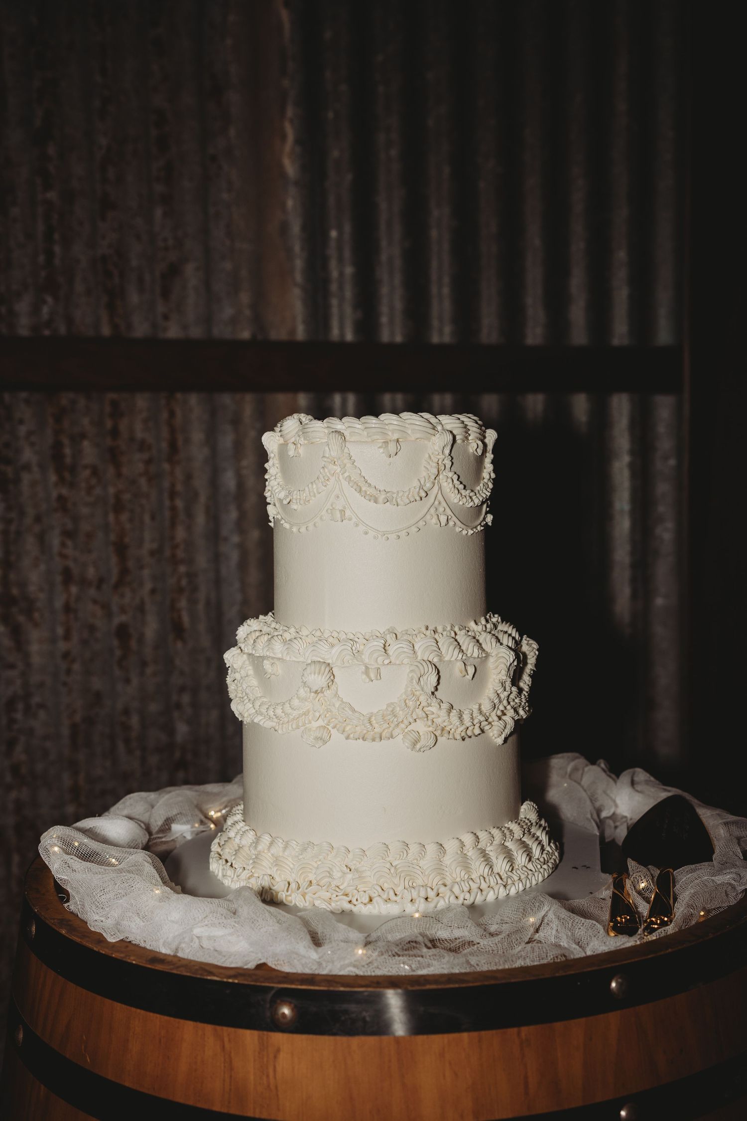 Elegant three-tiered white wedding cake with delicate lace piping details displayed on a wooden barrel base.