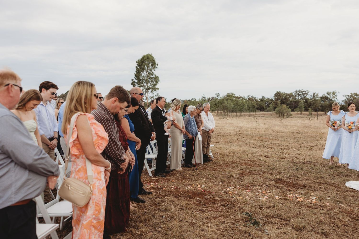 Wedding guests watching the ceremony from white chairs in an open field.