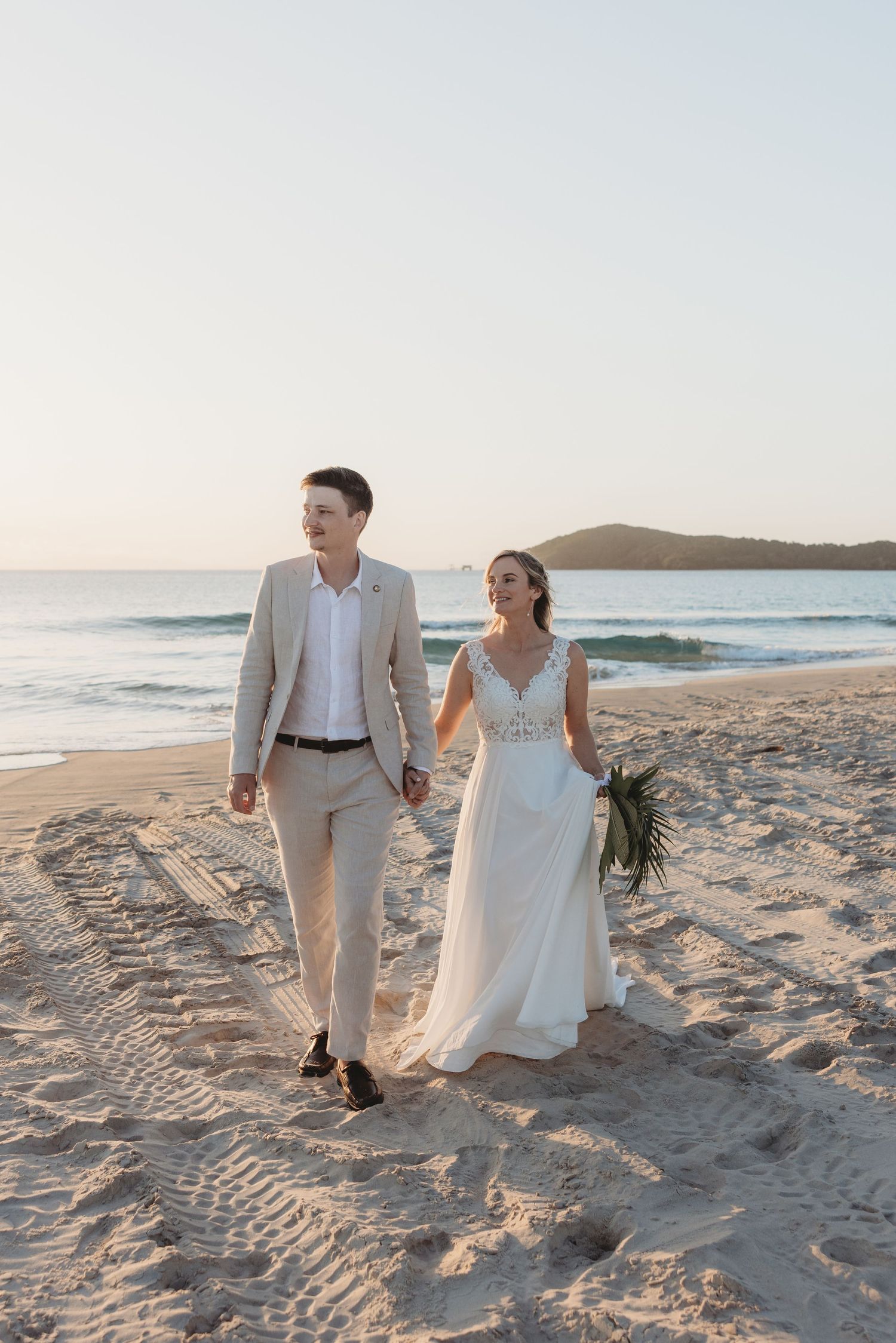 Newlyweds walking hand in hand along sandy beach during golden hour.