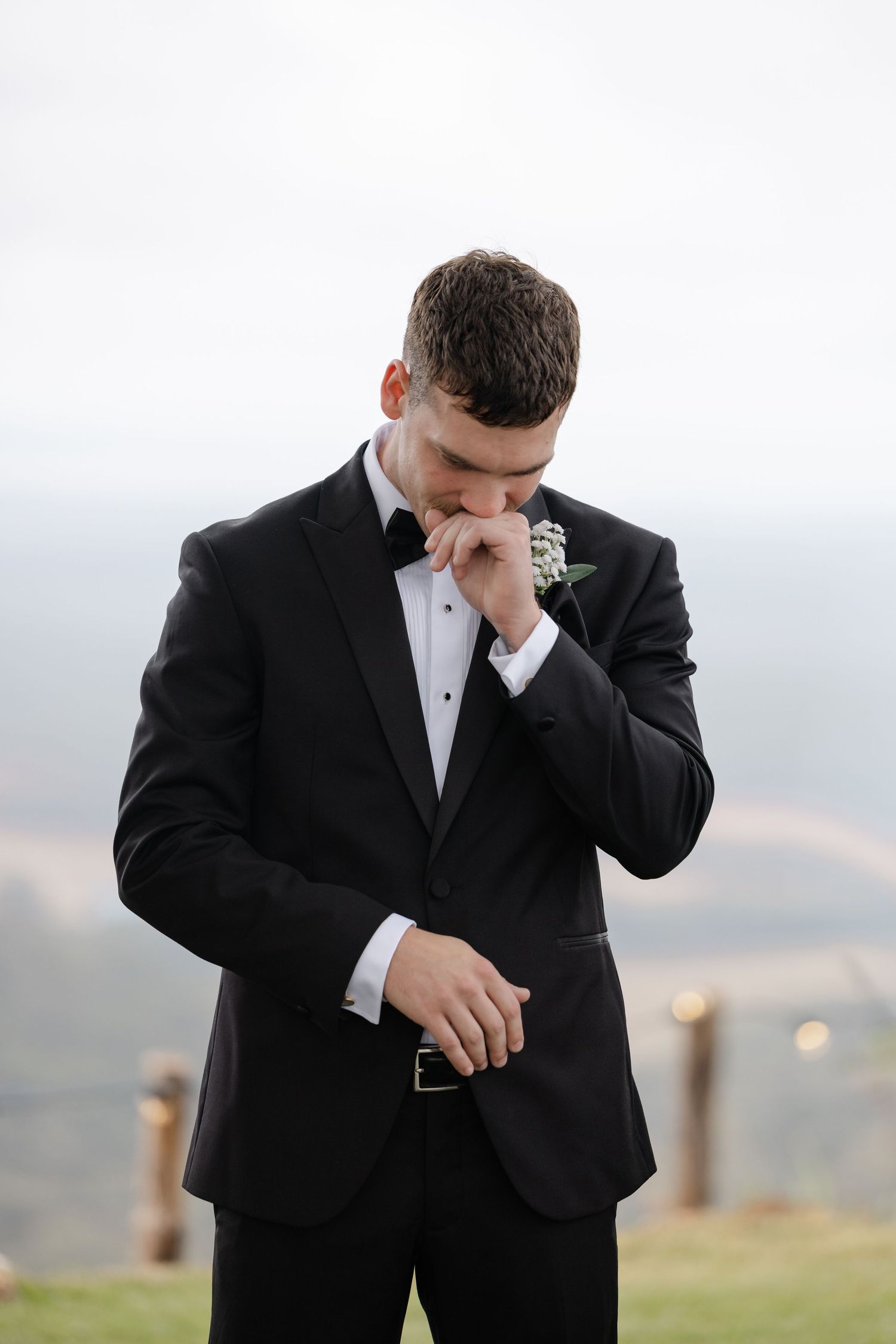 Emotional groom in black tuxedo with boutonniere looking down during outdoor wedding ceremony.