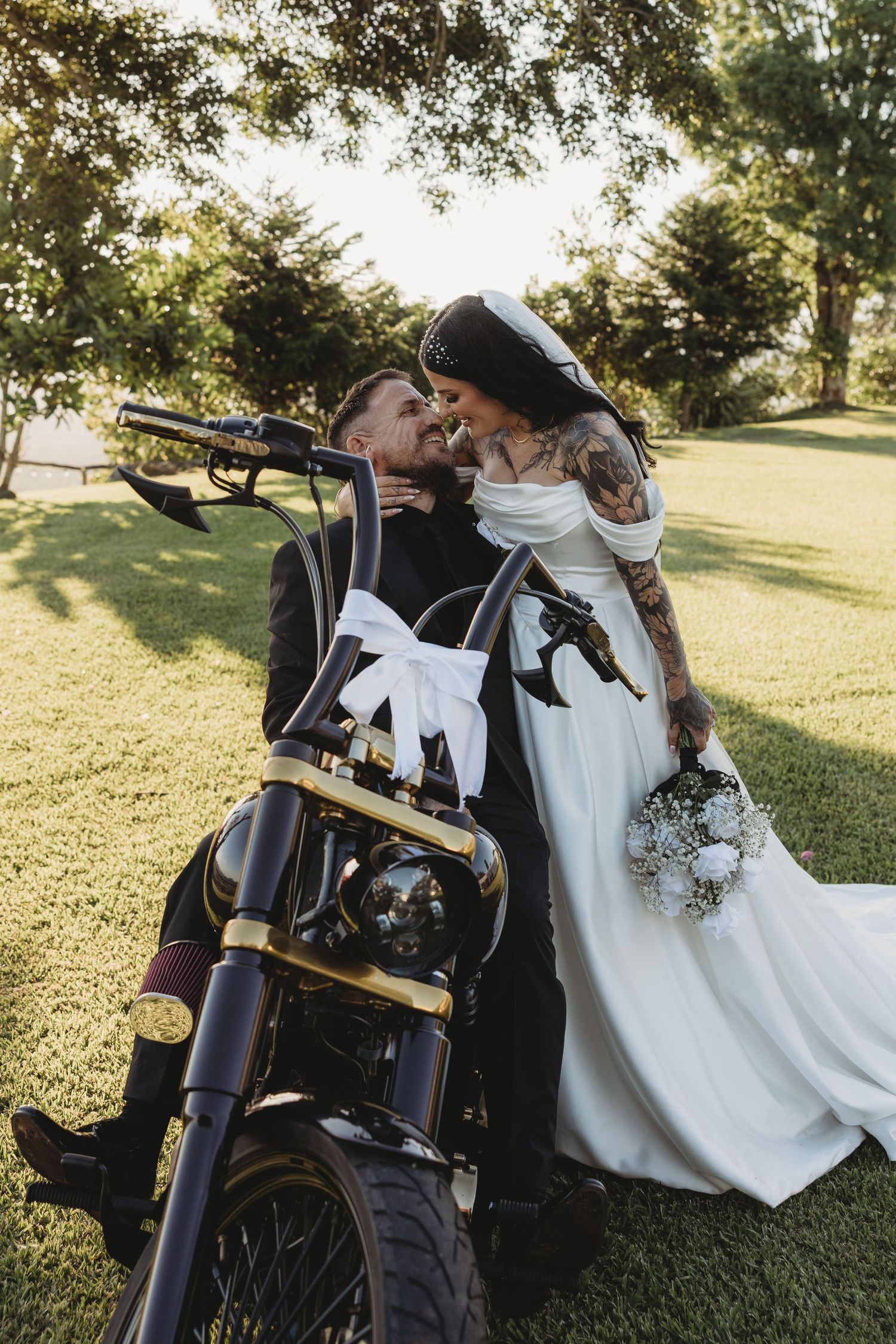 Couple posing with motorcycle in outdoor wedding photoshoot on sunny grassy field.