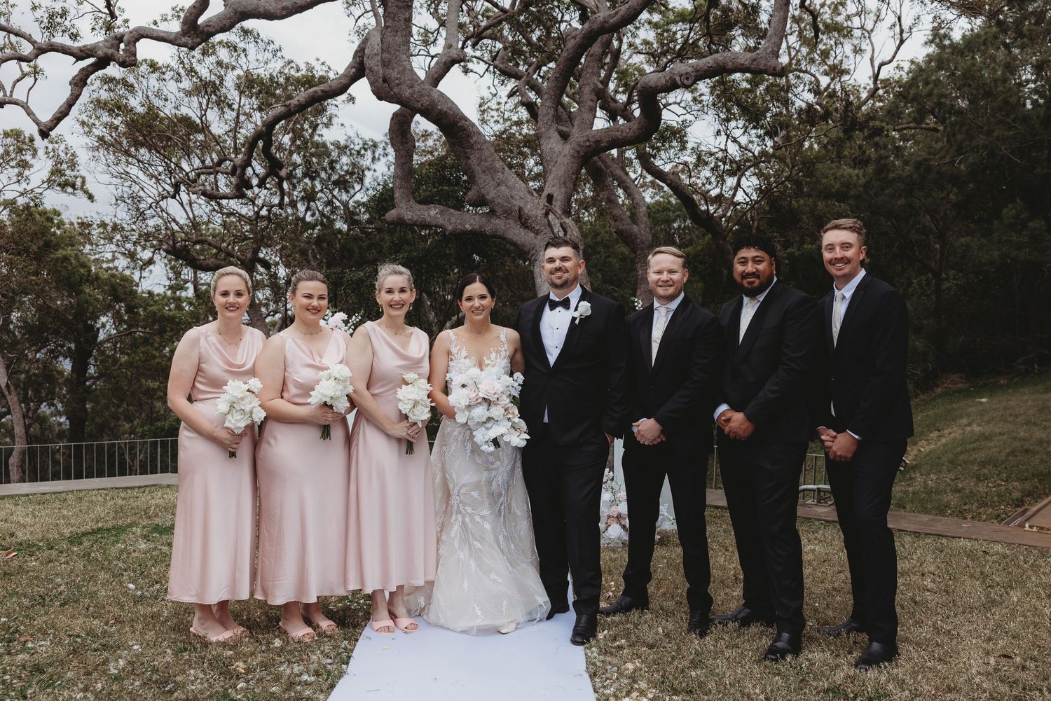 A wedding party poses for a formal photo outdoors with bridesmaids in pink dresses and groomsmen in black suits.