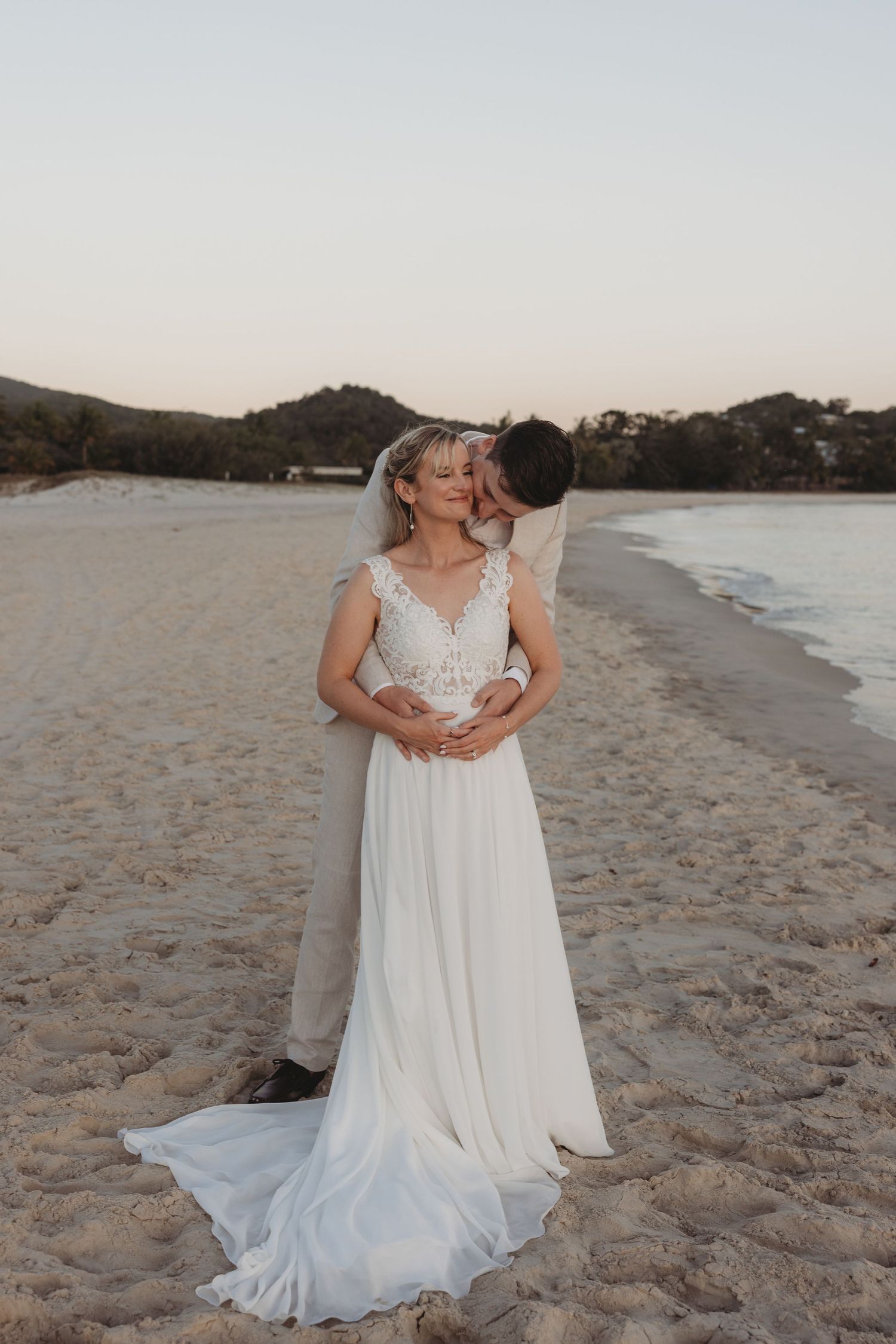 Bride in flowing white dress poses with partner on beach during golden hour.