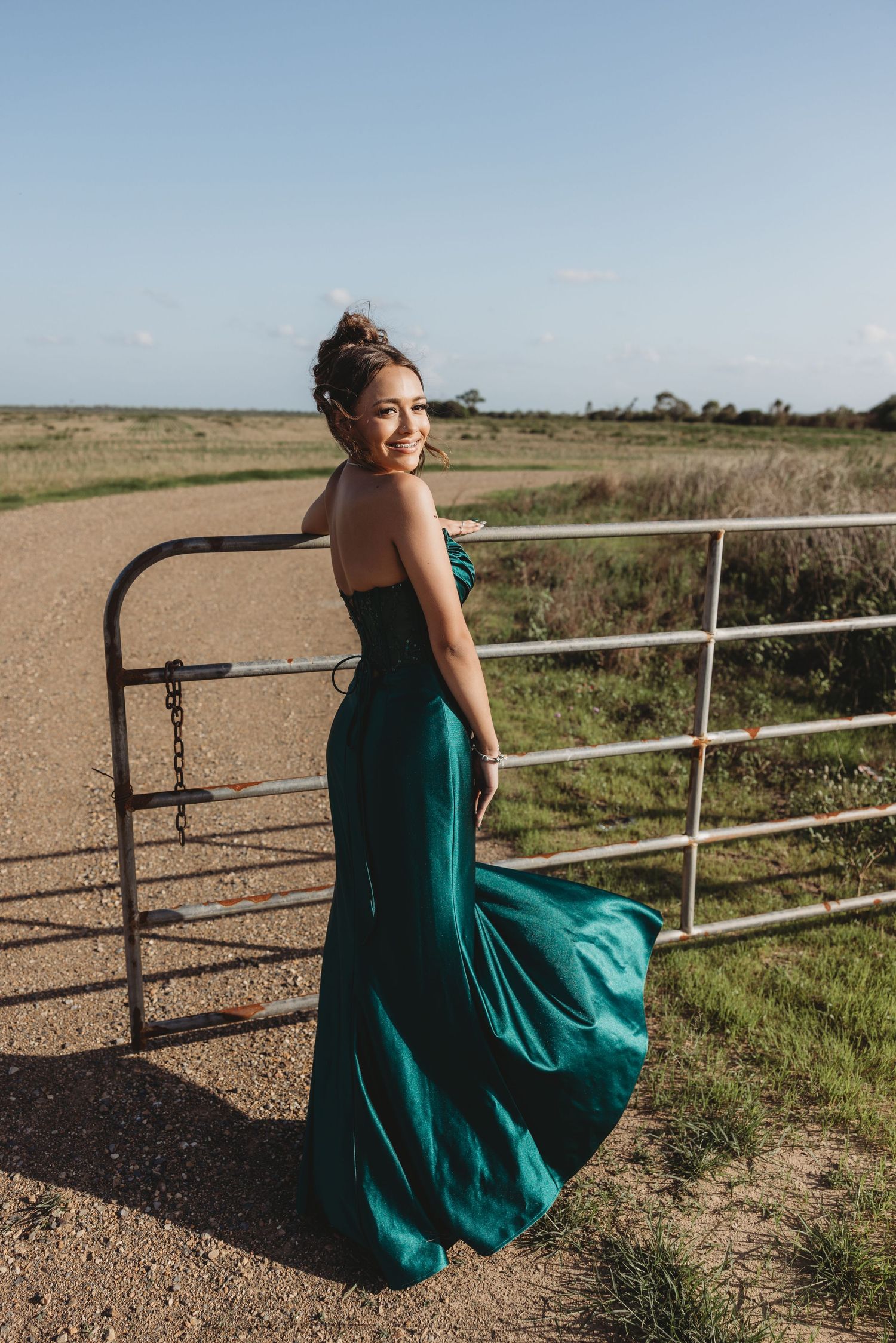 Woman in flowing emerald gown poses by rustic gate in countryside setting.