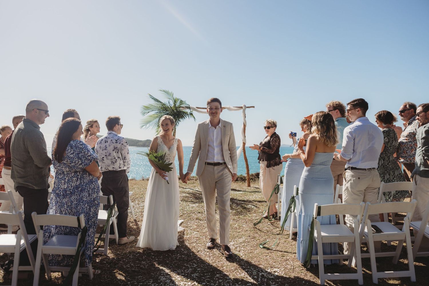 Wide shot of beach wedding ceremony with guests seated under bright blue sky.
