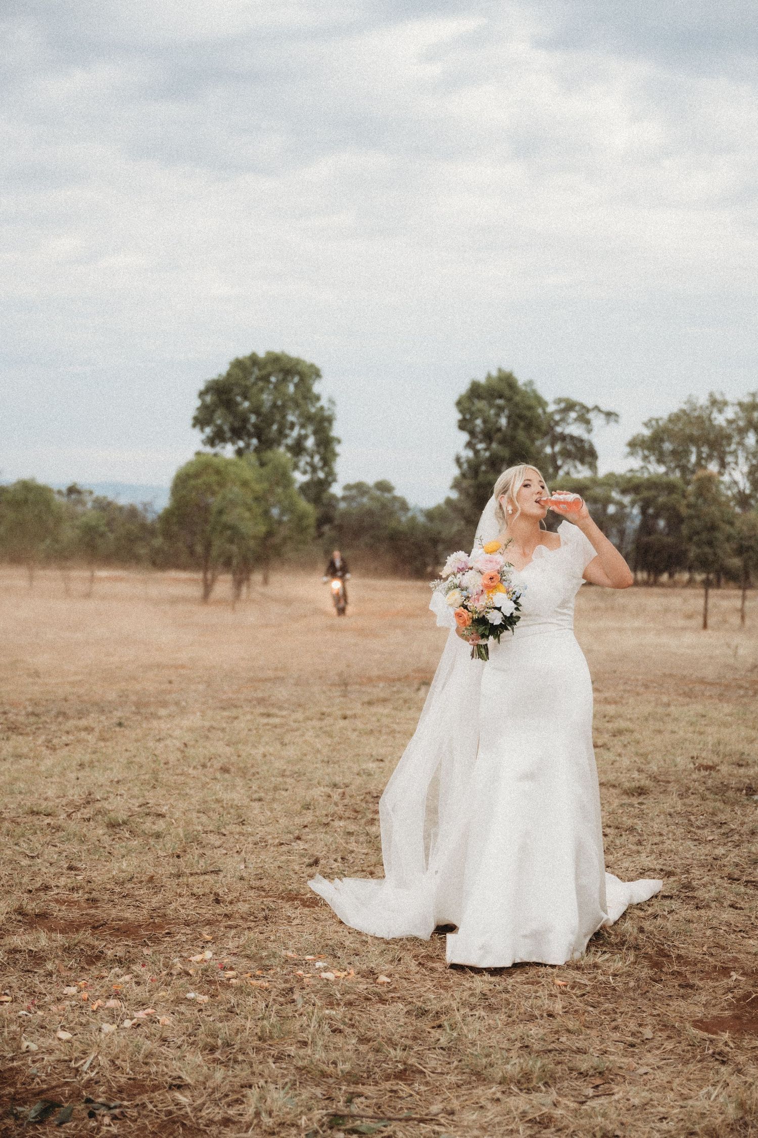 Bride in flowing white dress poses alone in rustic field with bouquet.