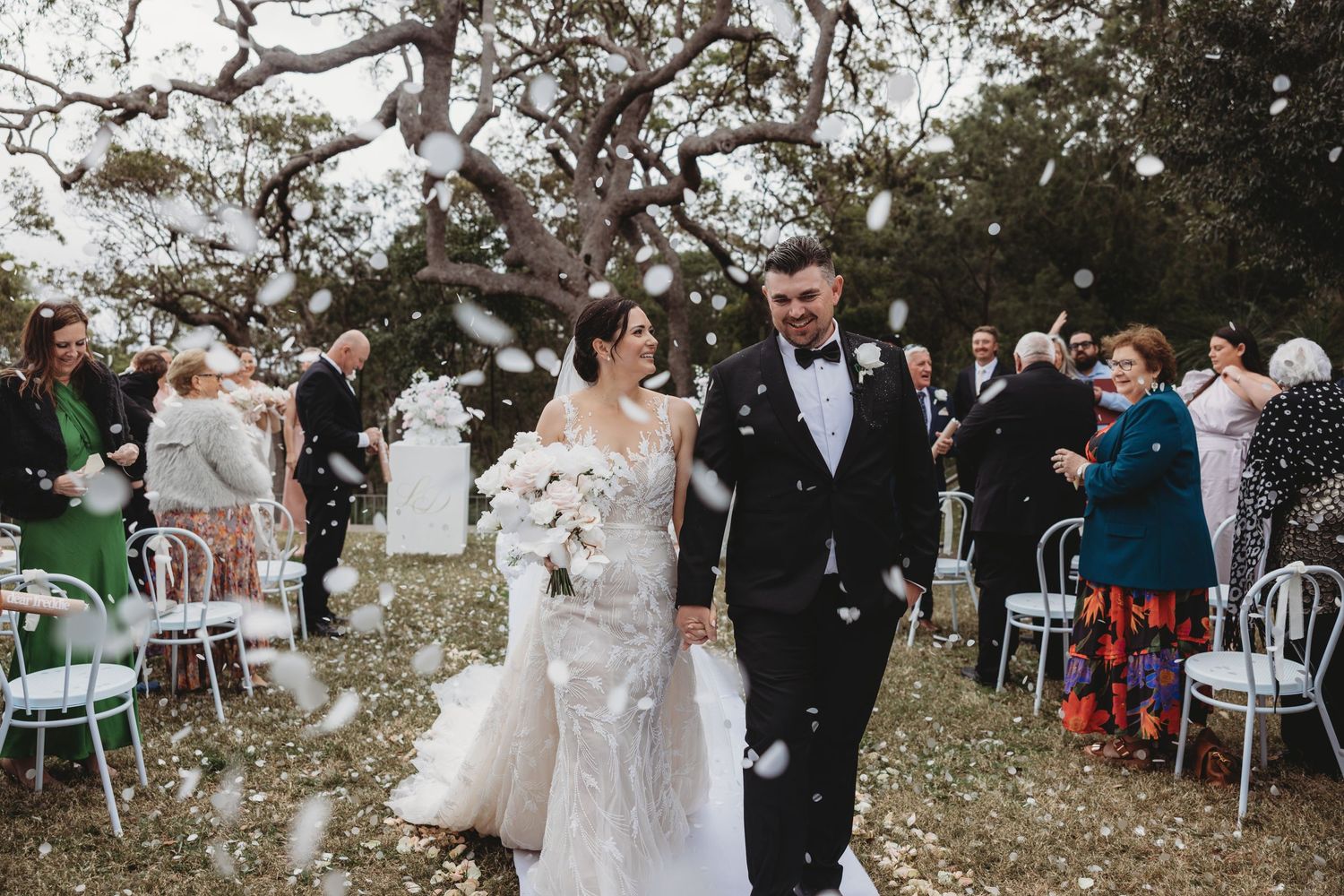 Wedding ceremony setup outdoors with white chairs and floral arrangements under trees.