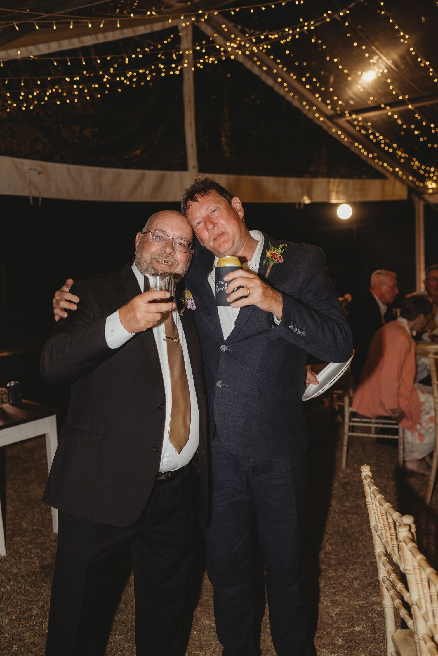 Two men in suits toasting drinks at an outdoor wedding reception under fairy lights at night.