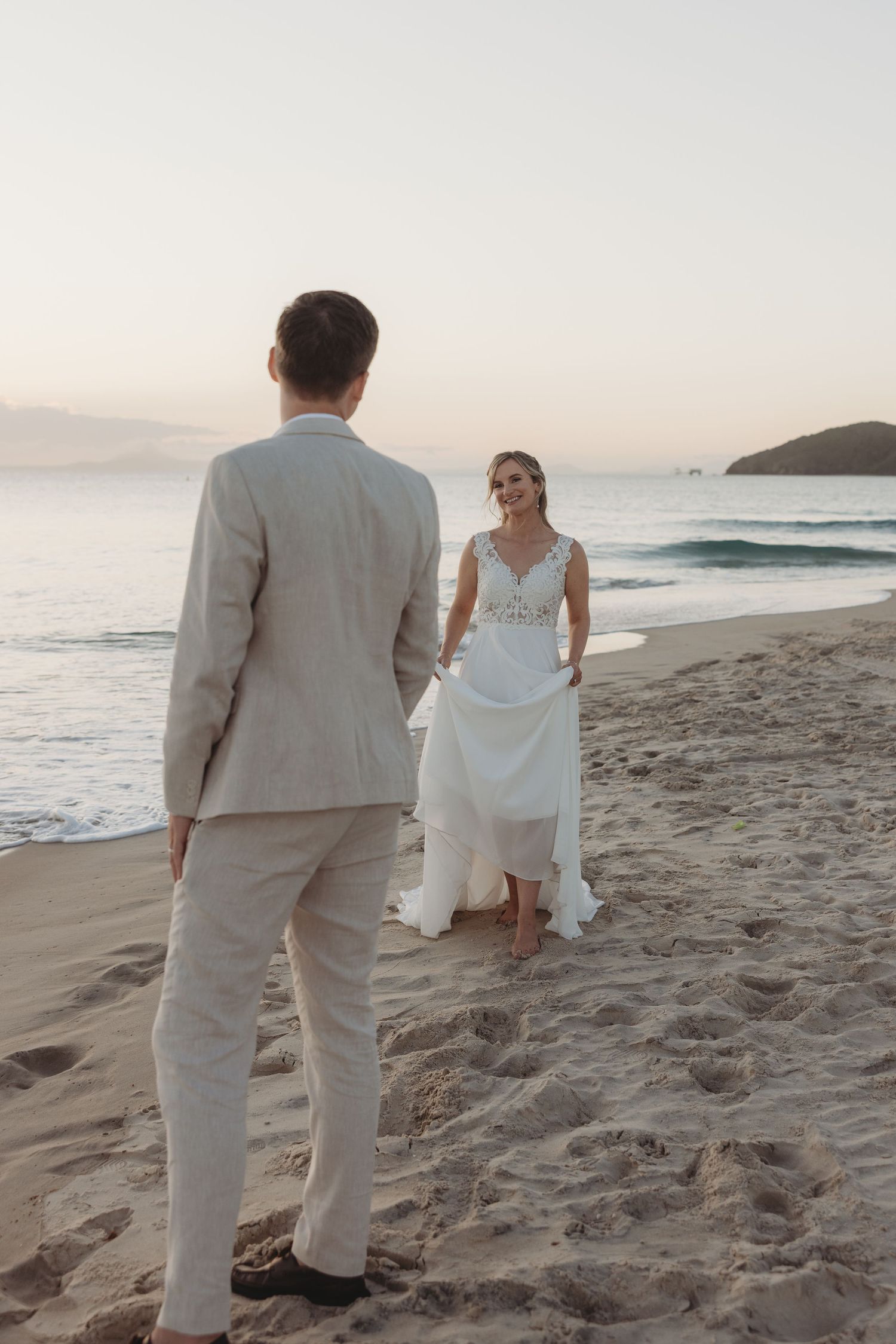 Couple walks along sandy beach at sunset wearing light colored formal attire.