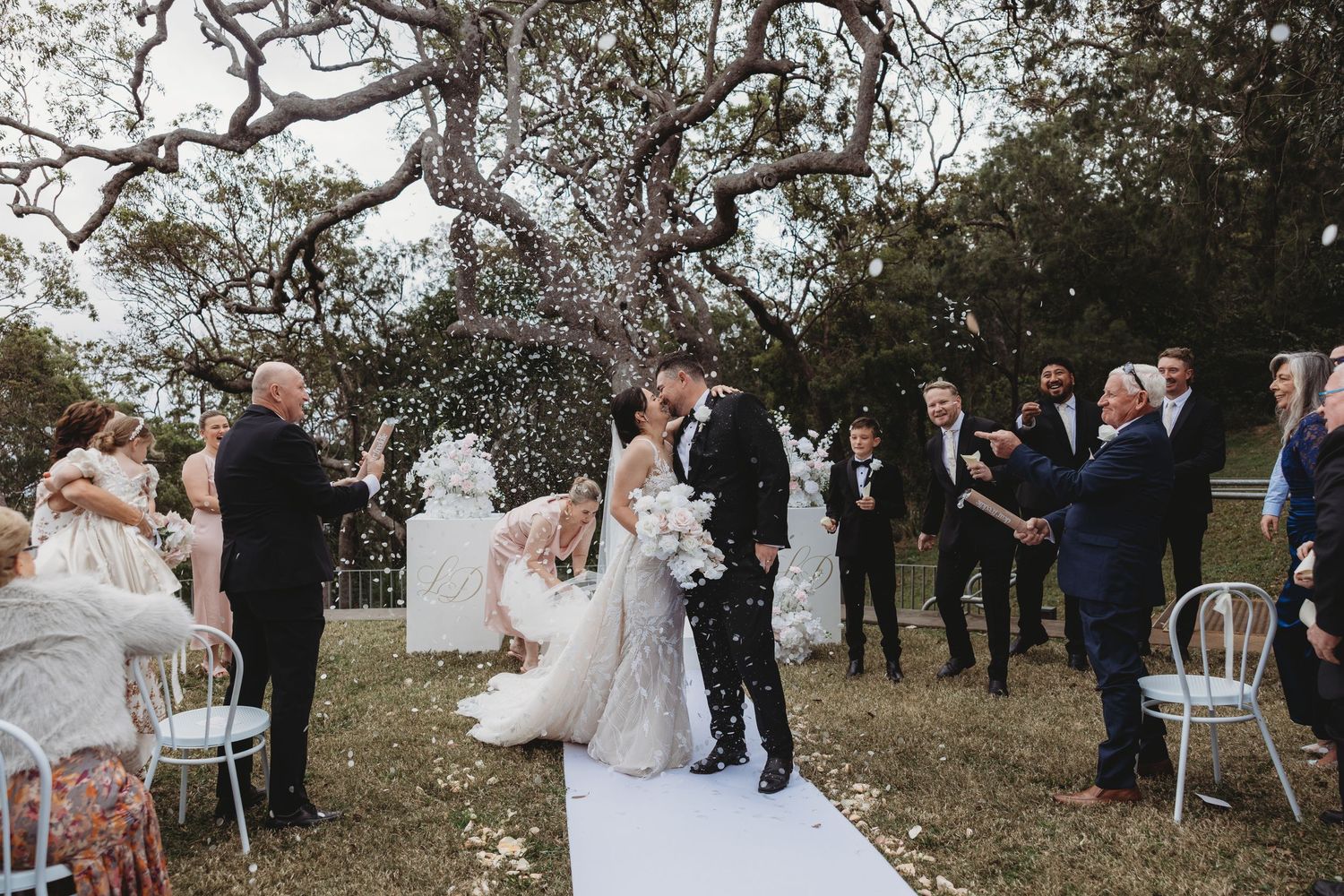 Wedding guests celebrate with confetti during an outdoor ceremony under trees.