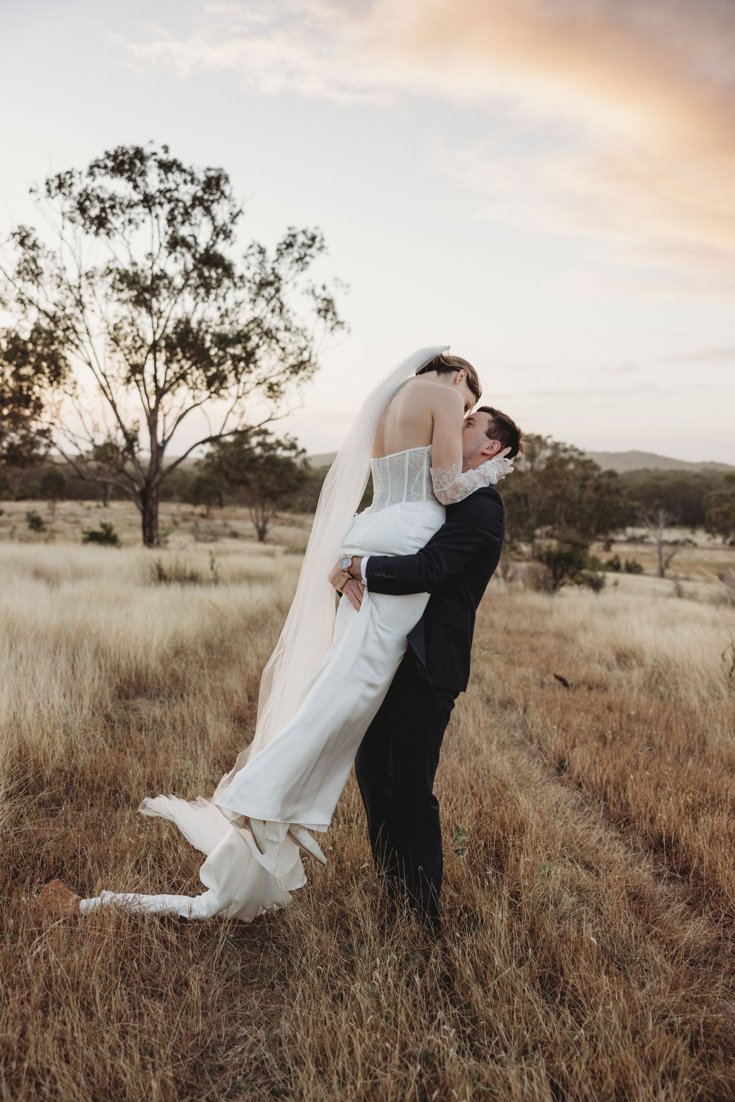 Groom lifting bride in white gown and veil in a golden field at sunset during wedding portraits.