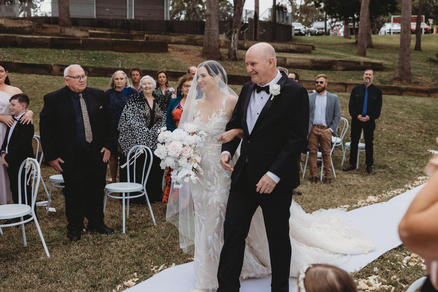 An outdoor wedding ceremony setup with white chairs arranged in rows under tall trees on a grassy area.
