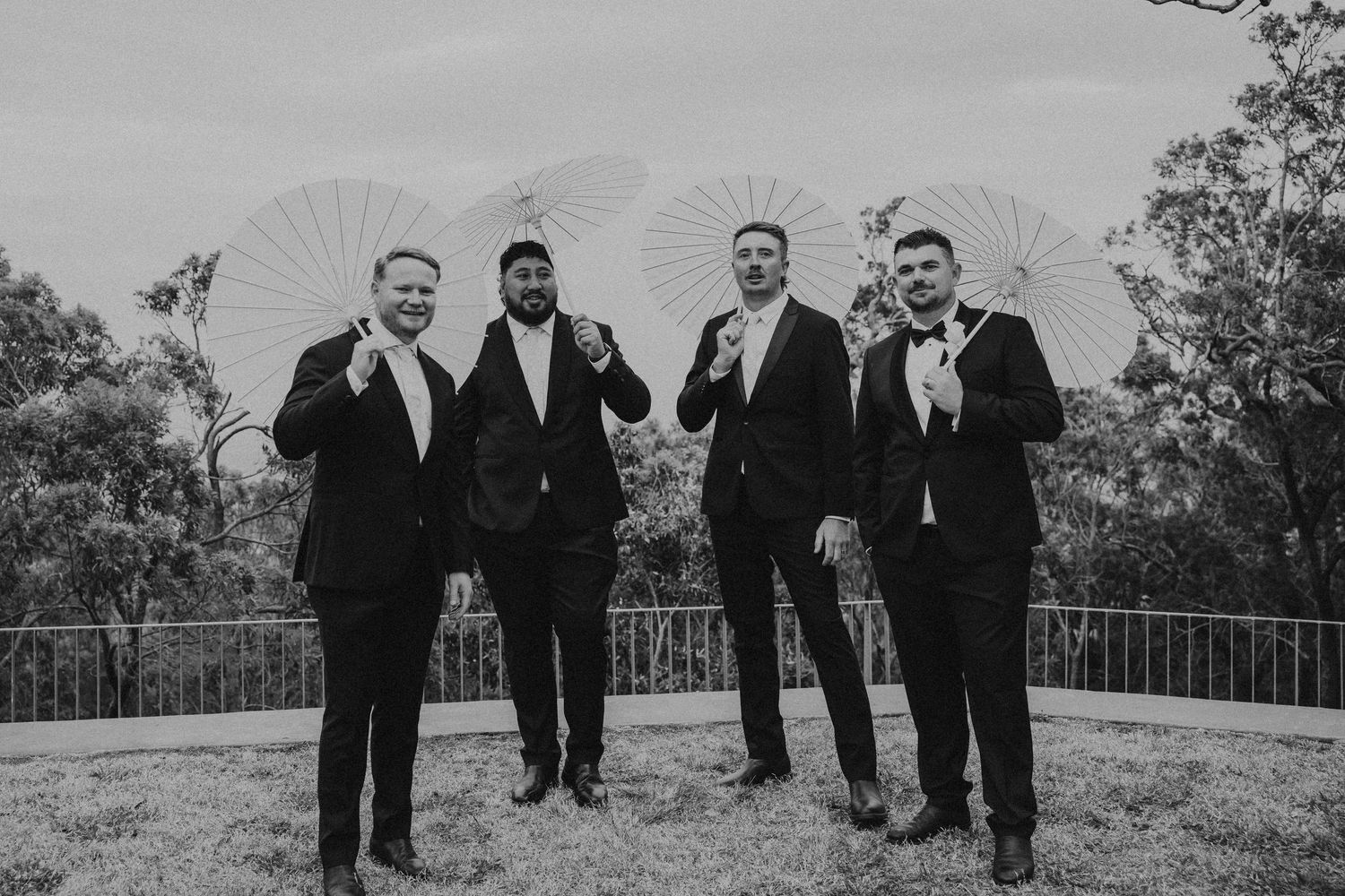 Groomsmen in formal black tuxedos stand together outdoors for wedding photos.