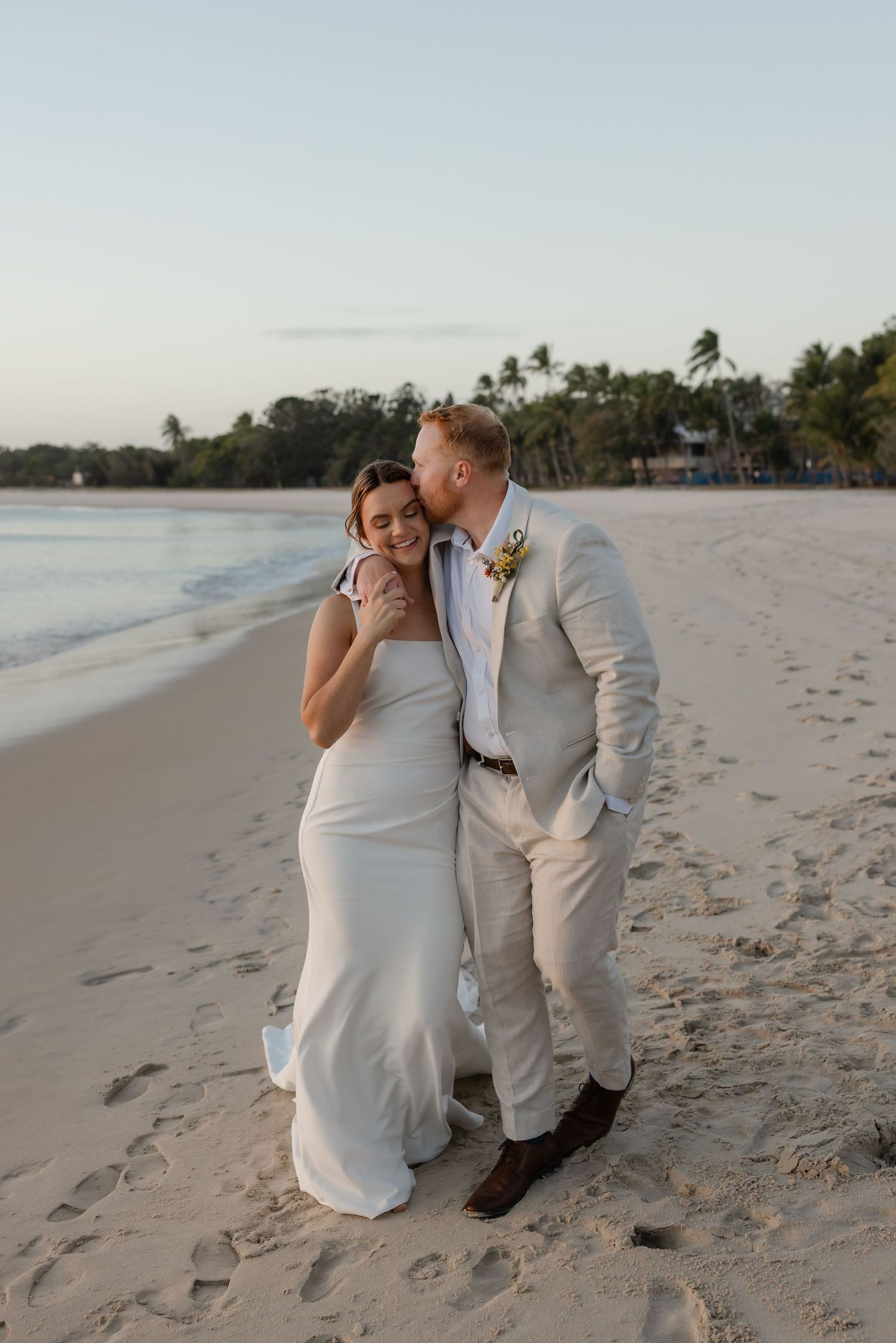 A bride and groom share a romantic kiss on a sandy beach at sunset, dressed in a white gown and tan suit.