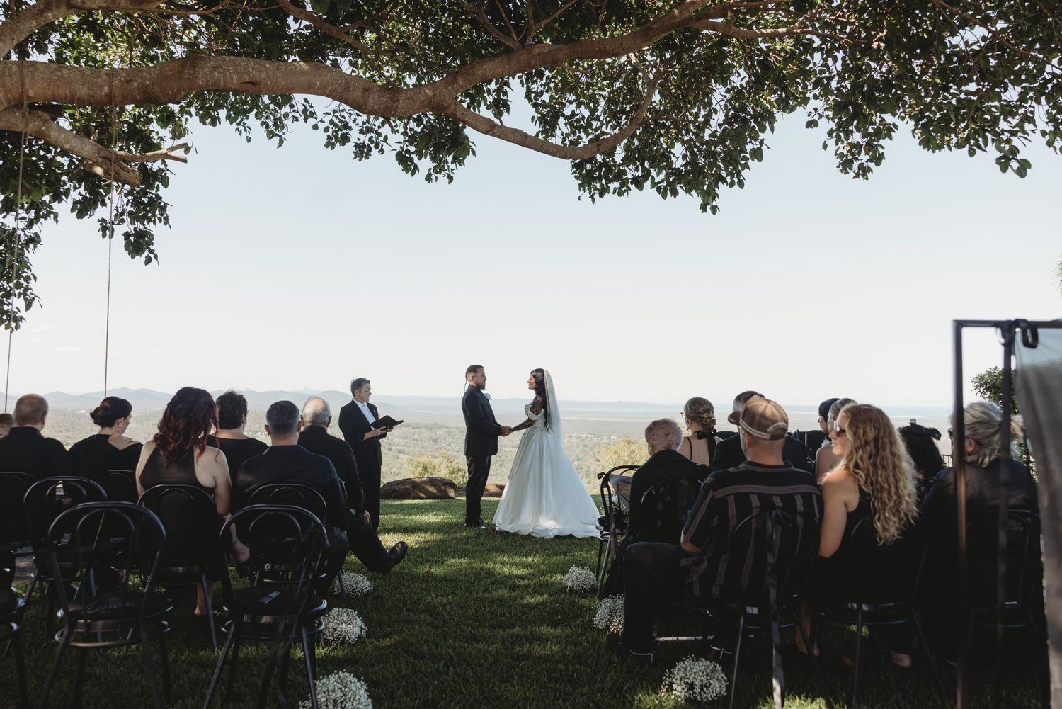Outdoor wedding ceremony on a grassy cliff overlooking the ocean with guests seated under a tree canopy.