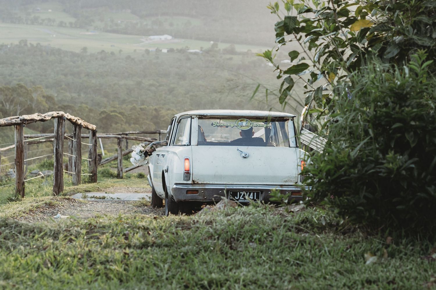 Scenic outdoor wedding venue with rolling hills and a vintage car visible in the distance.