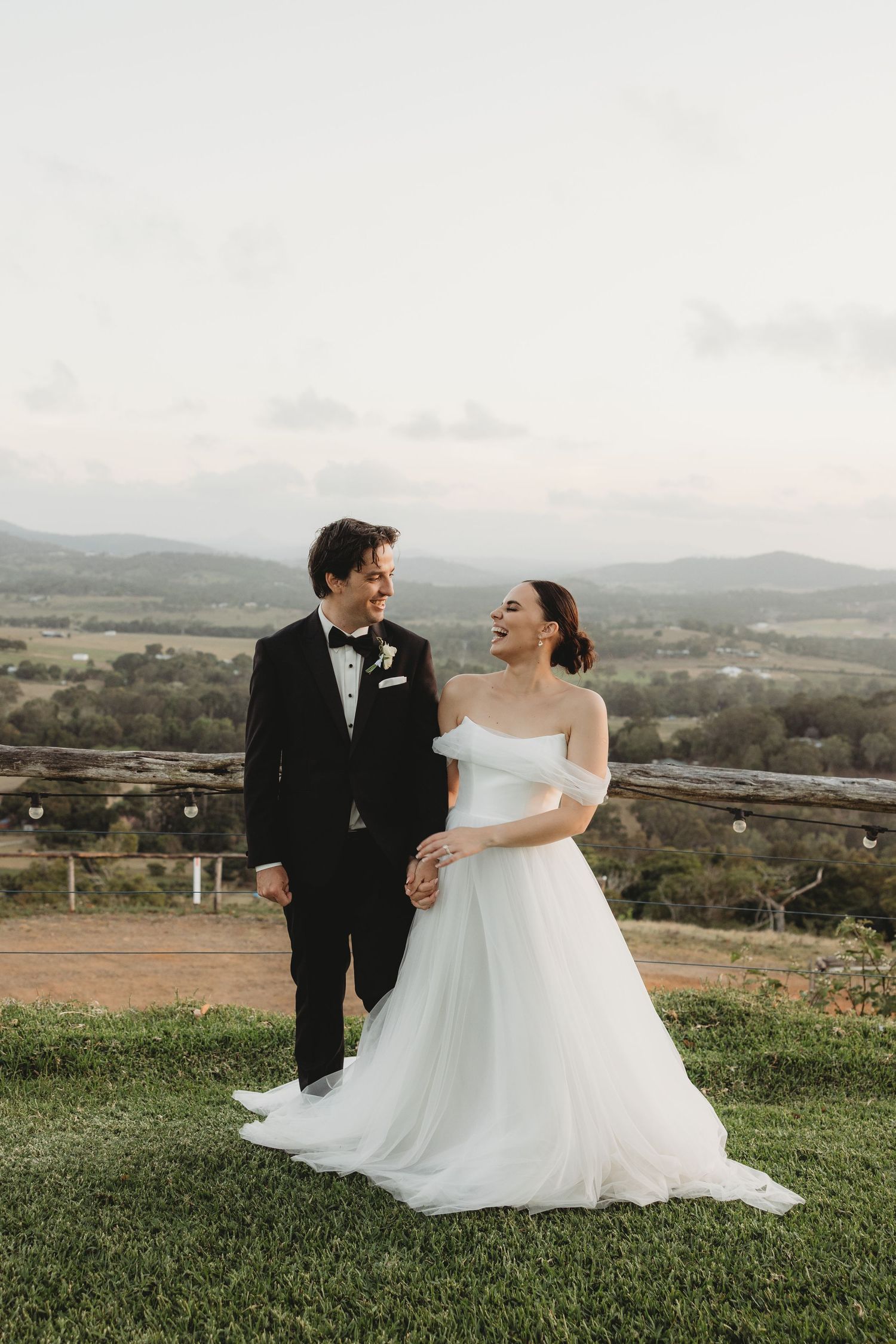 A joyful wedding couple laughing together on a scenic overlook with rolling hills in the background.