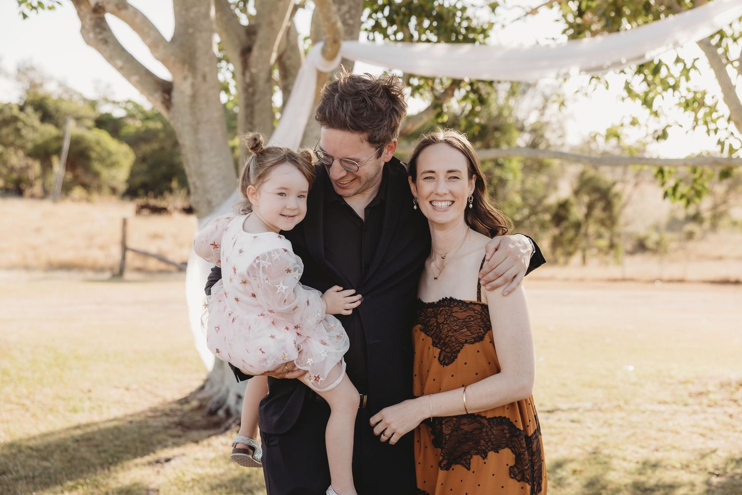 Happy family of three smiling together outdoors under large tree at a wedding celebration.