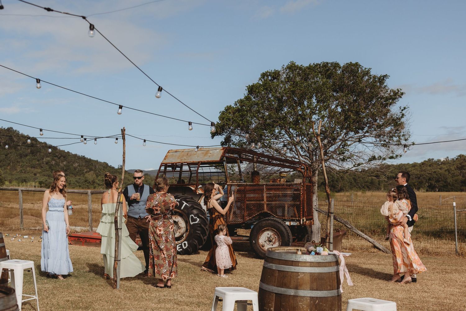 Wedding guests mingle outdoors near vintage truck with string lights at rustic farm venue.