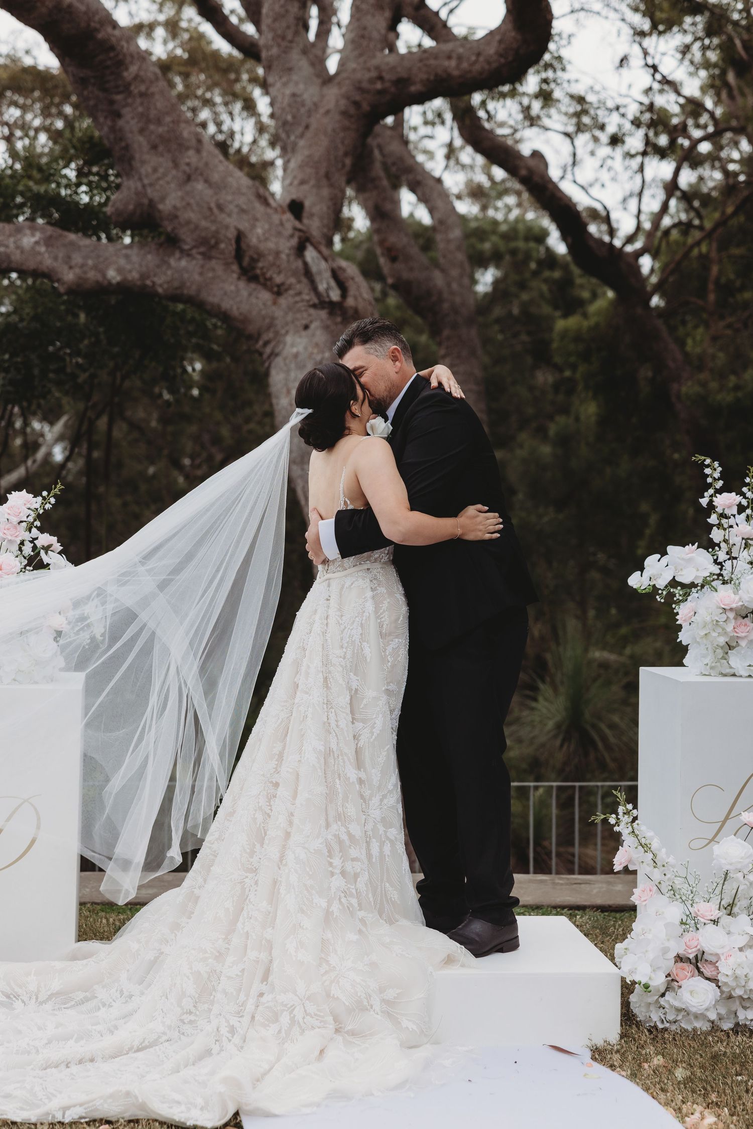 A romantic wedding moment captured outdoors with a flowing white veil and floral arch decorations.