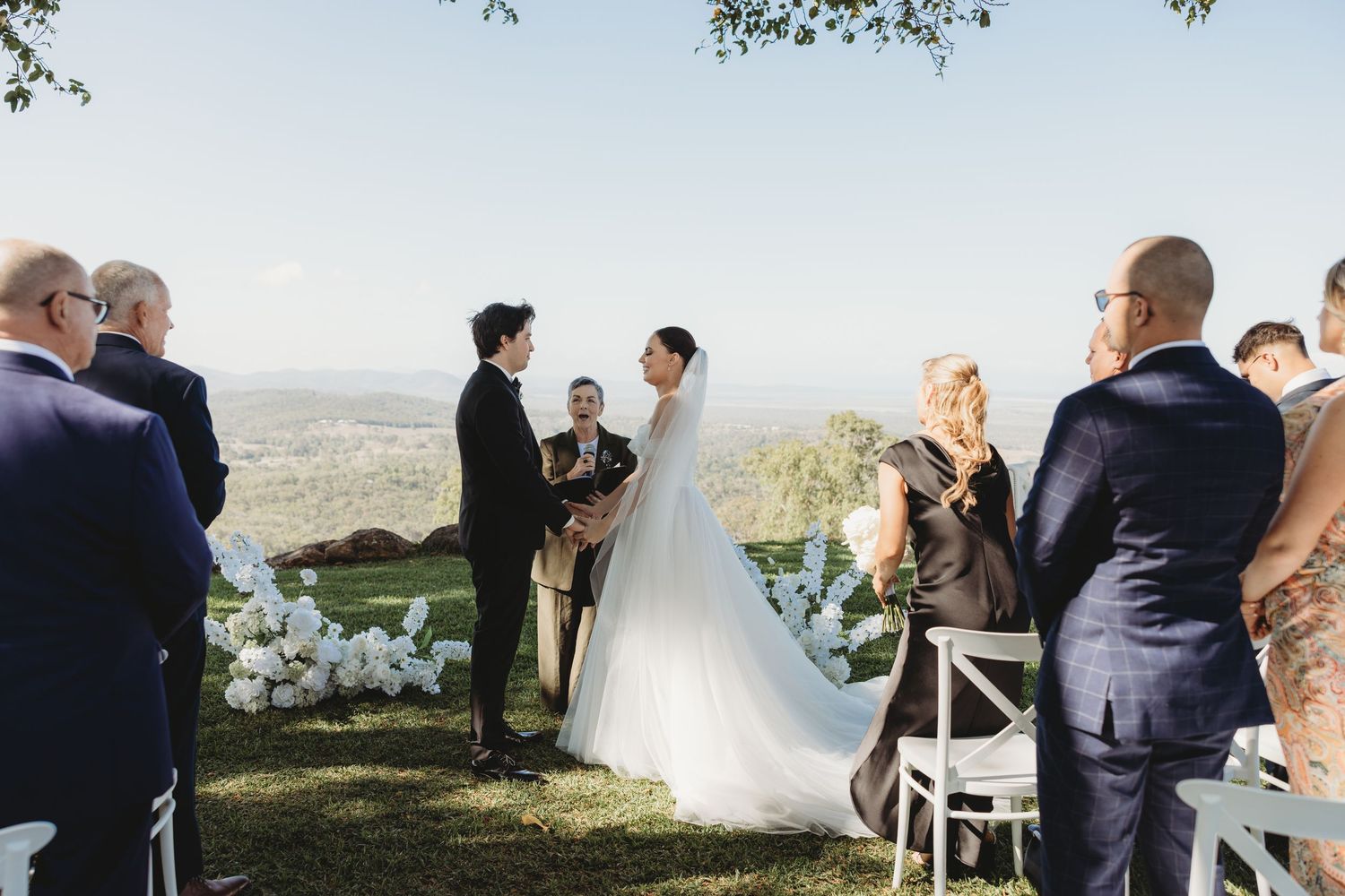 Wedding ceremony in progress on a hilltop with guests seated in white cross-back chairs under blue skies.