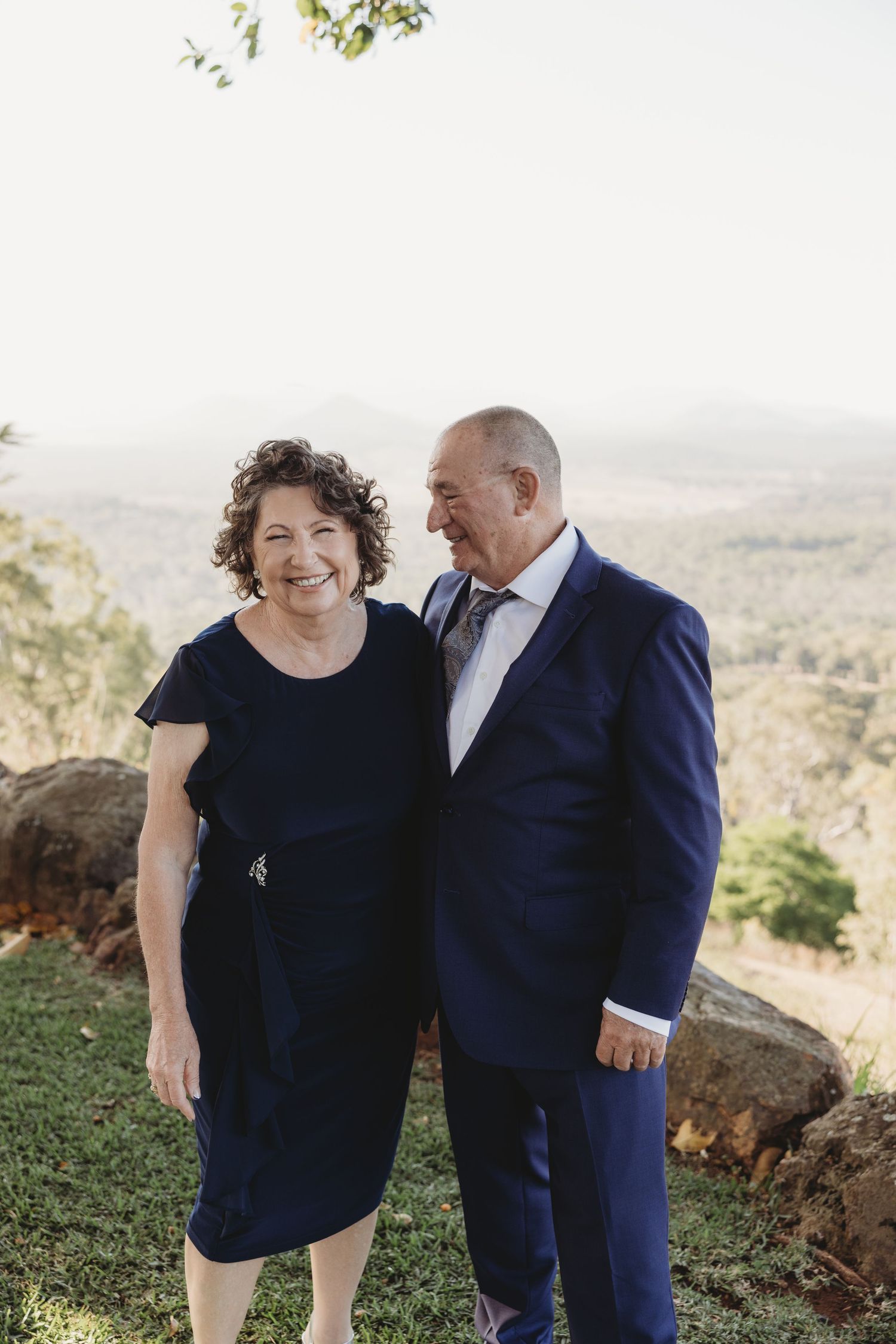 A couple in navy blue attire embracing on a hilltop with a scenic valley view.