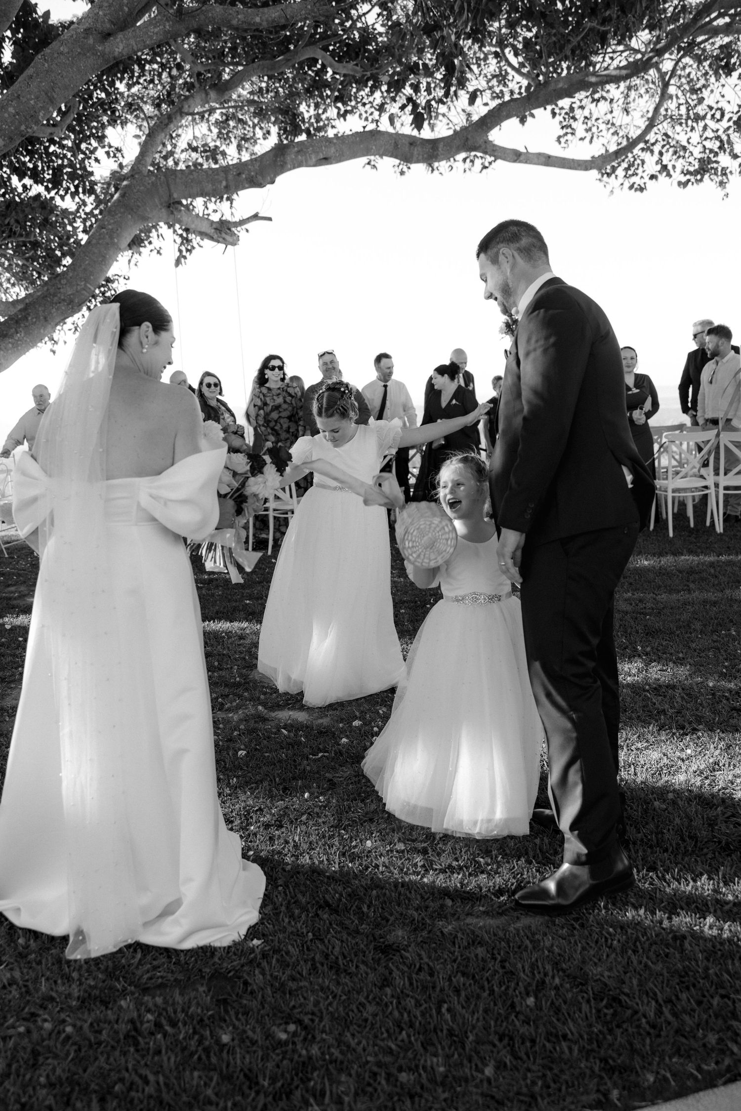 Black and white photo of bride and groom with flower girls celebrating at an outdoor wedding ceremony.