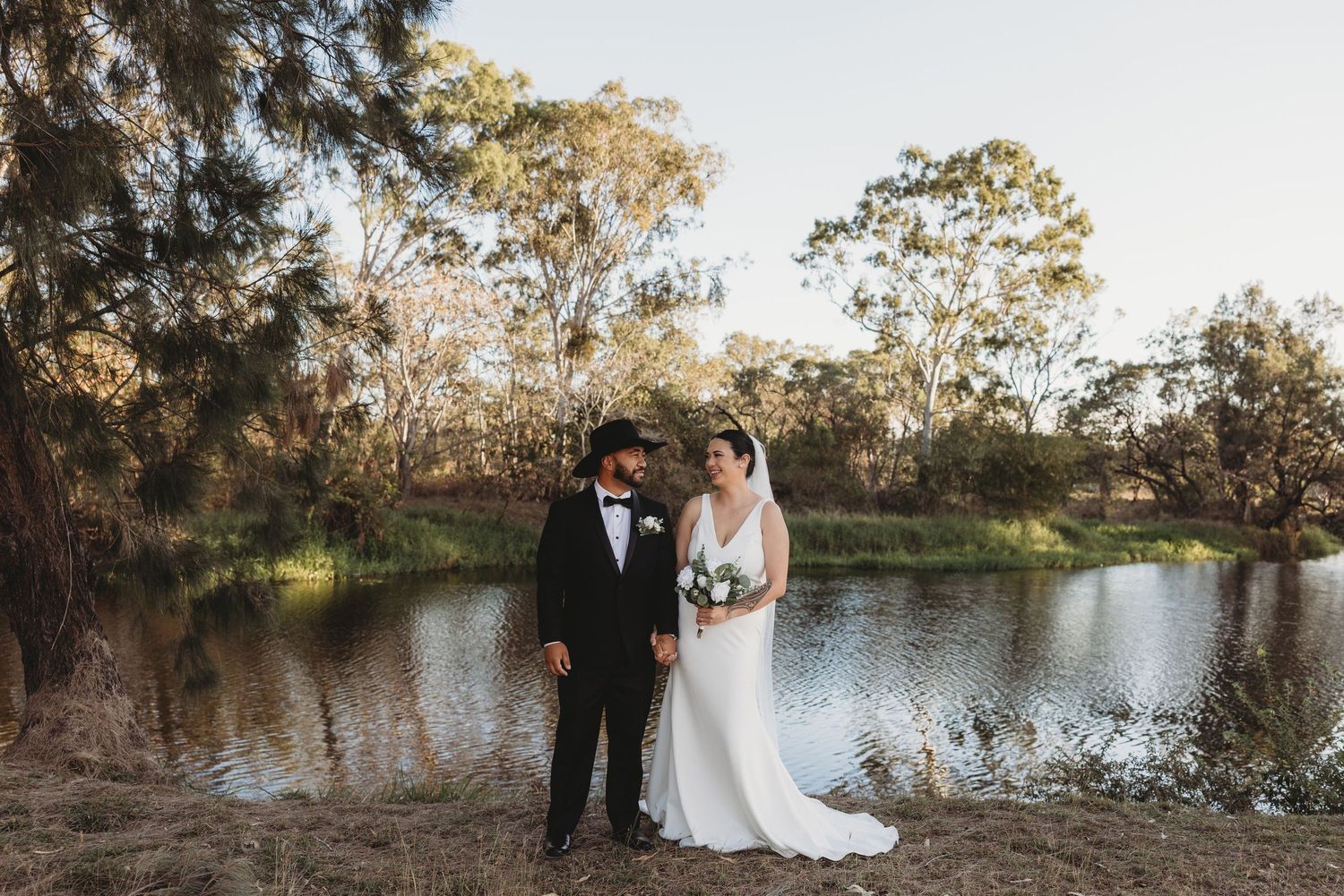 A wedding couple stands together by a serene lake surrounded by trees at dusk.