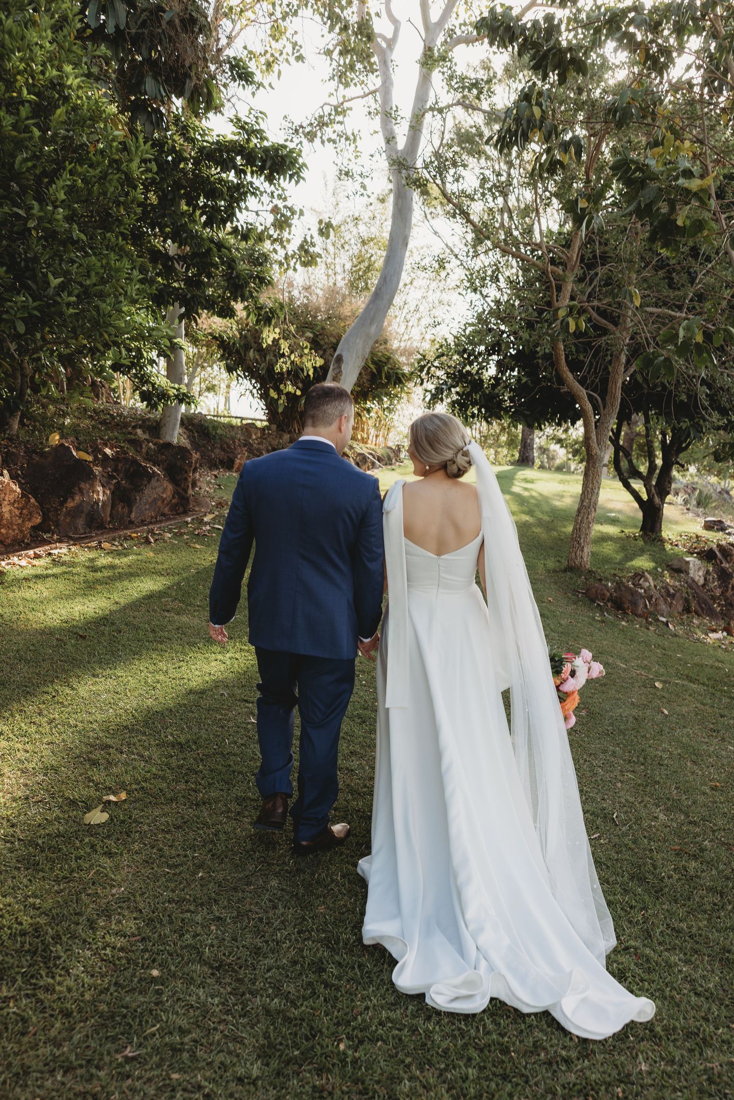 A wedding couple walks hand in hand through a garden setting with flowing white dress and navy suit.