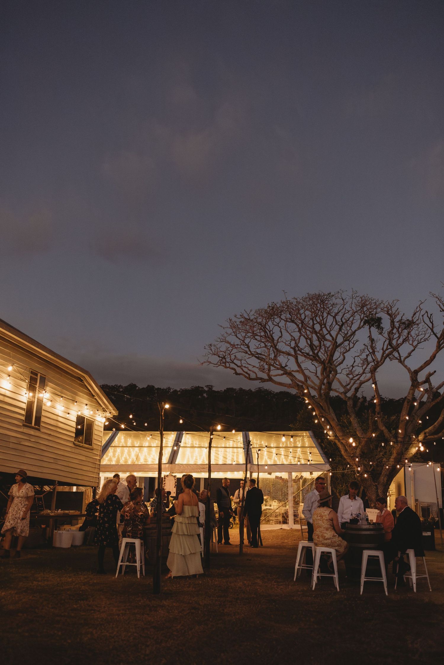 Wedding guests mingle under string lights at an outdoor evening reception beside a rustic country venue.