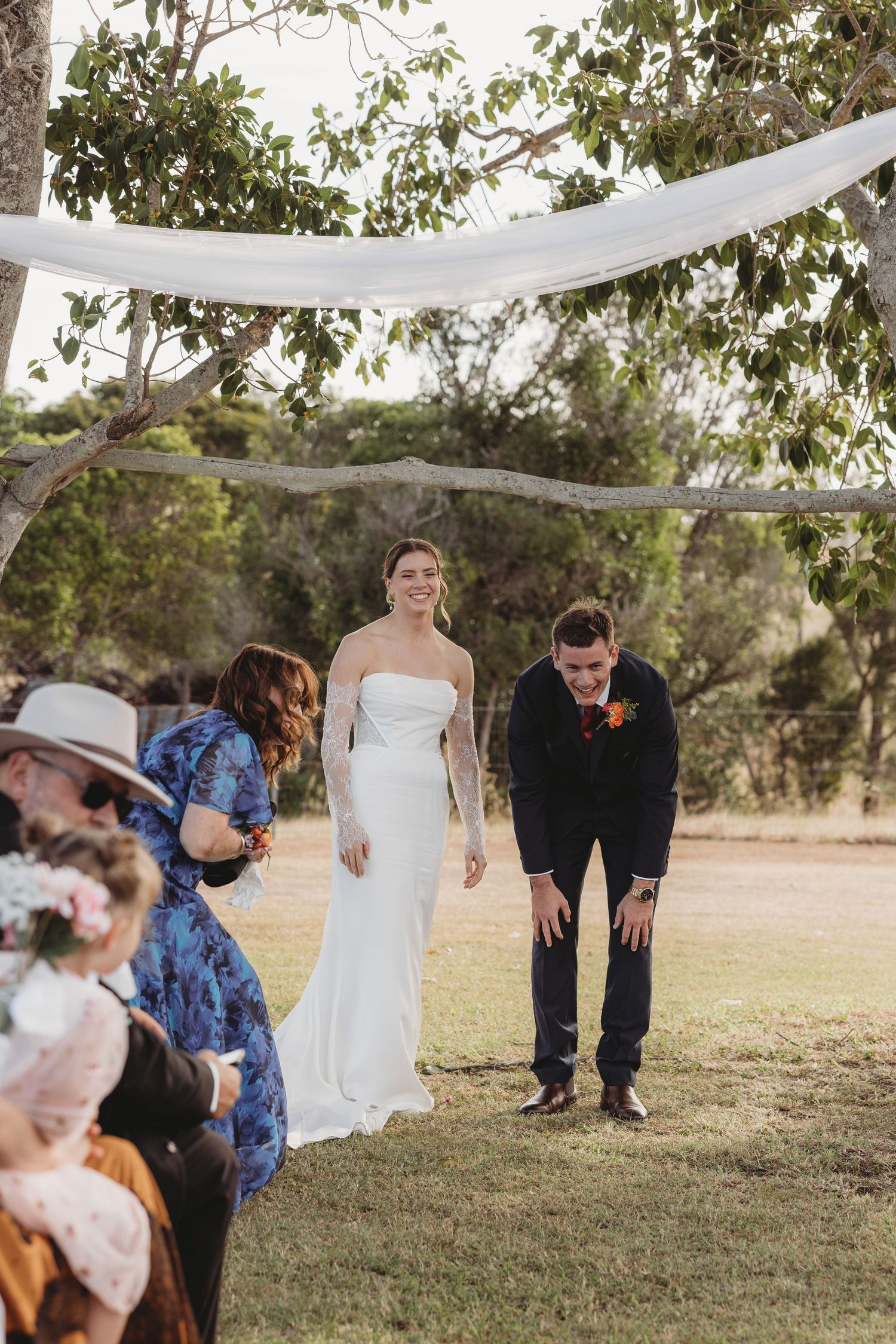 Smiling bride and groom walk down the aisle at an outdoor wedding ceremony under a draped tree canopy.
